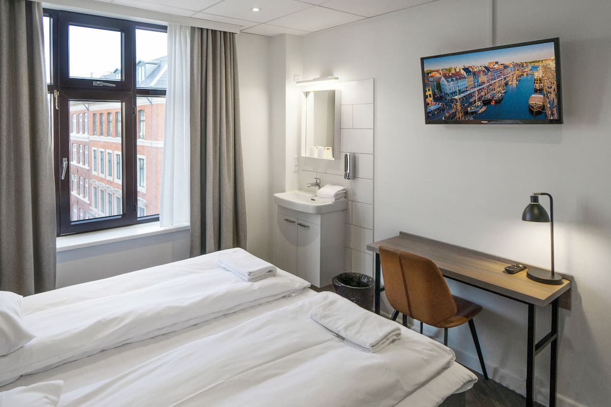 Une chambre d’hôtel équipée d’un lit, d’un bureau, d’une chaise, d’un lavabo et d’une grande fenêtre avec des rideaux.