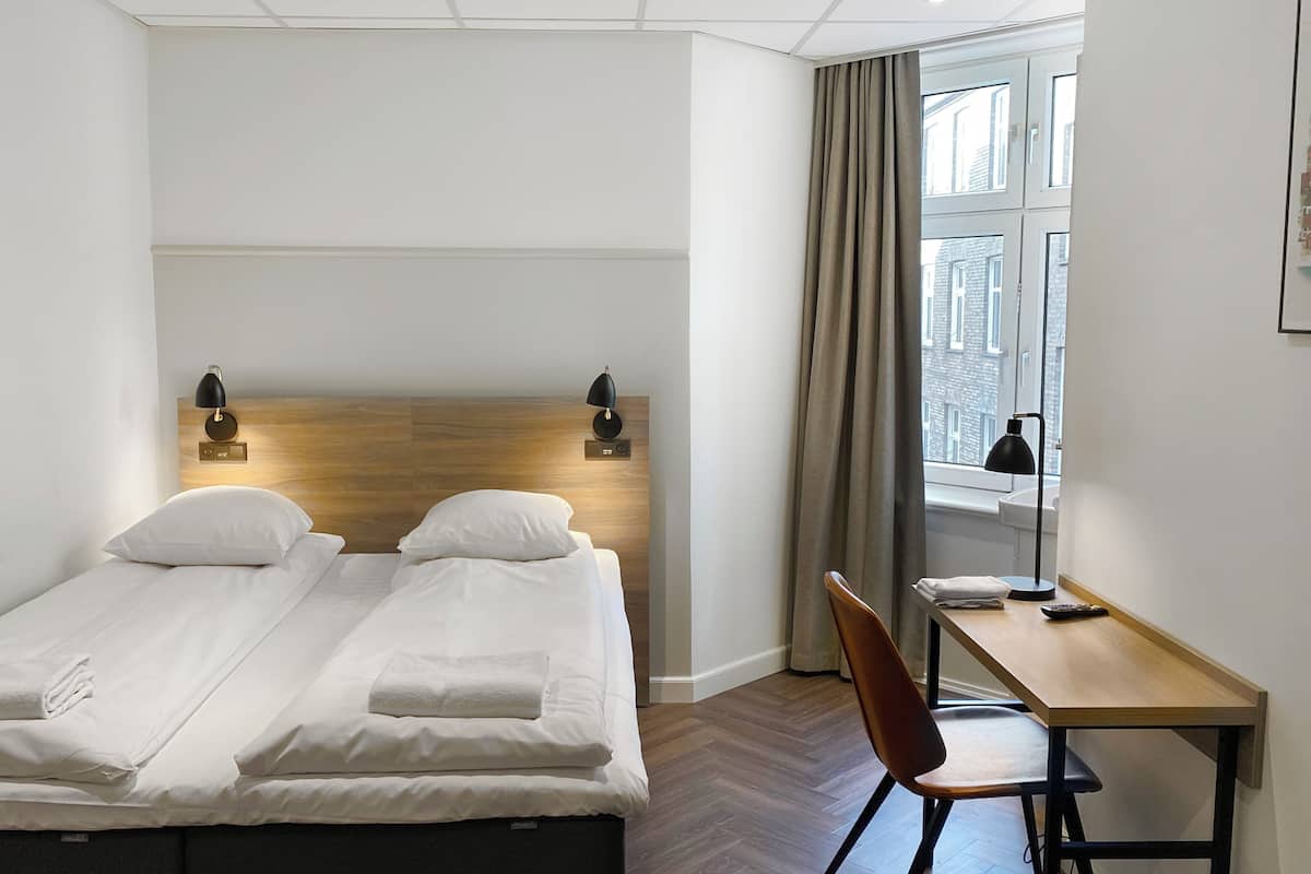 Une chambre d’hôtel avec deux lits simples, une tête de lit en bois, un bureau avec une chaise et une fenêtre avec des rideaux.