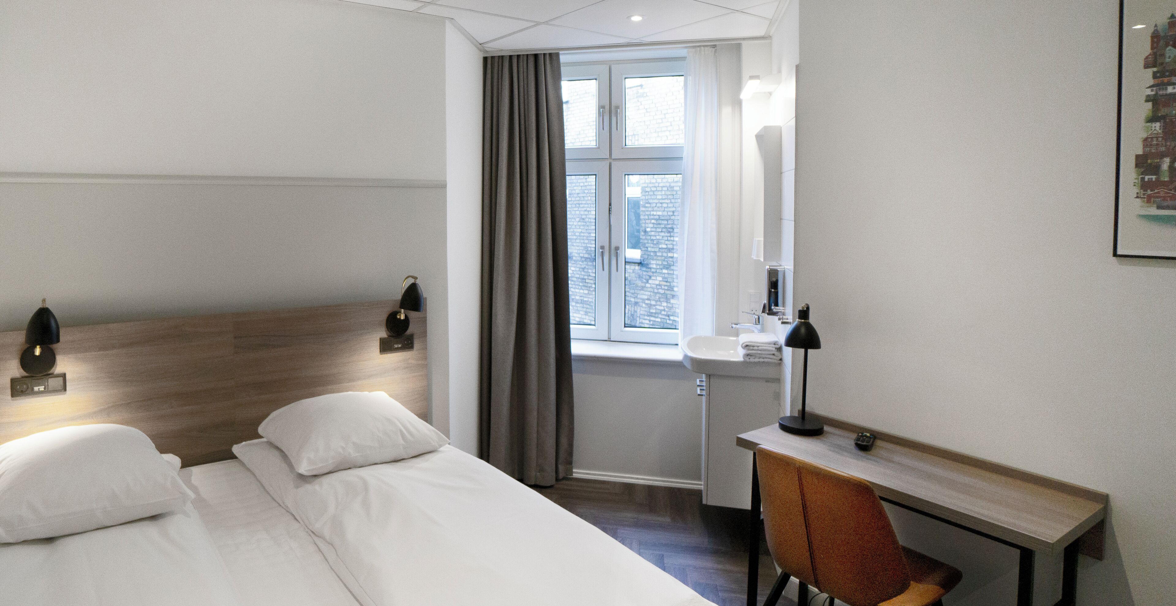 A hotel room with a bed, a desk, a chair, a window with curtains, and a wall-mounted lamp.
