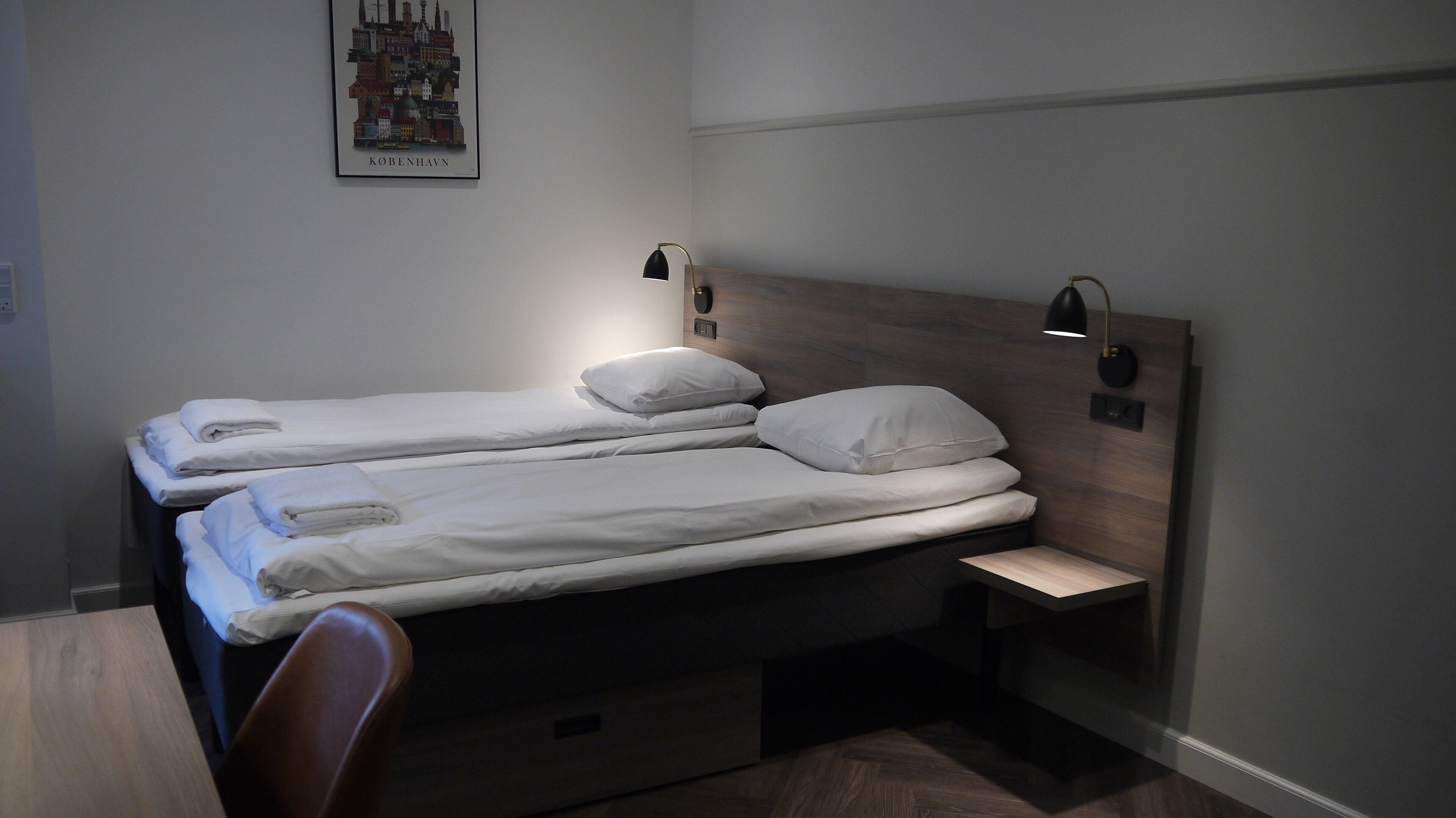 A hotel room with two single beds, a wooden headboard, bedside lamps, a small table, and a chair.
