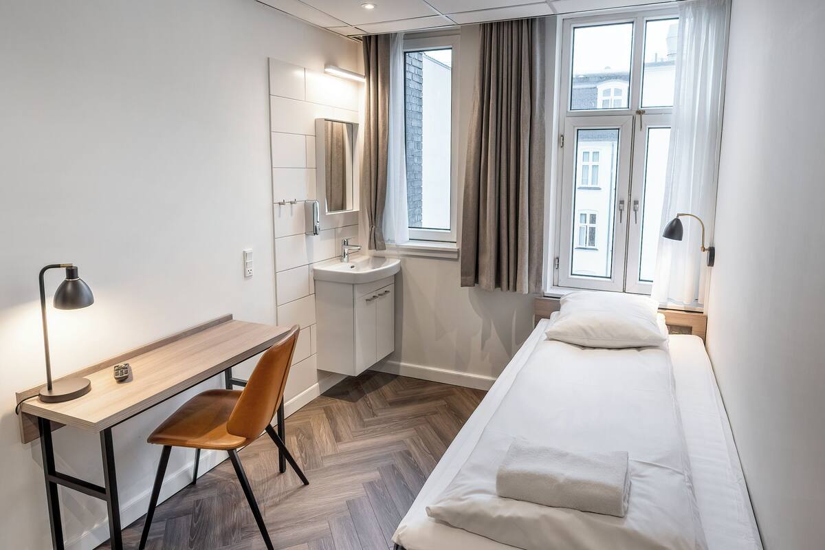 Une chambre d’hôtel moderne avec un bureau en bois, une chaise, un lit avec du linge de lit blanc, une petite salle de bain avec un lavabo et un miroir, et une fenêtre avec des rideaux.