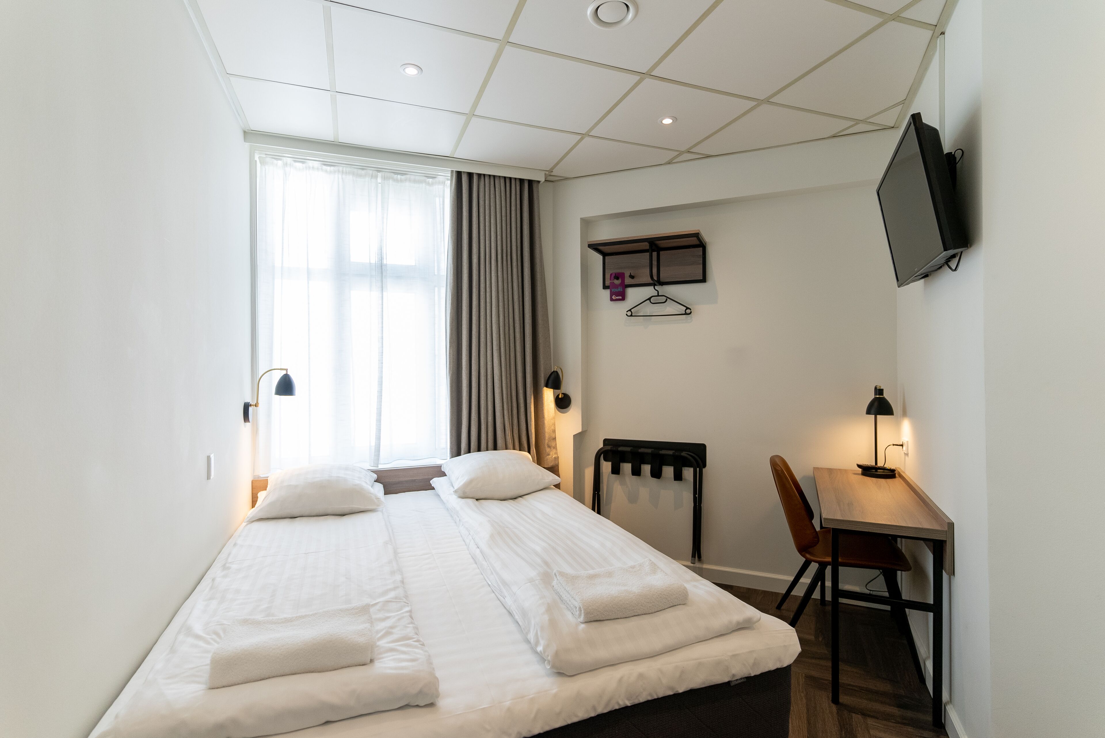 A hotel room with a bed, a desk, a chair, a TV, and a window with curtains.