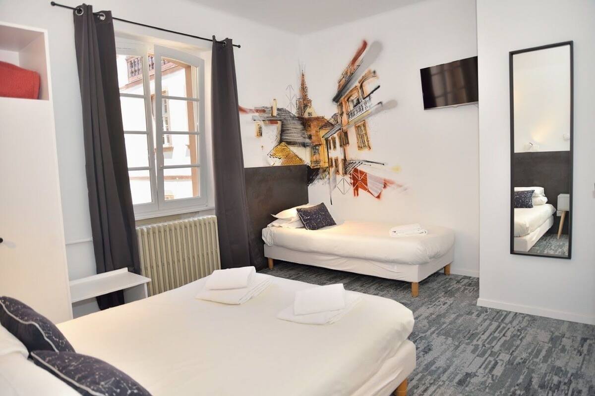 A hotel room with a bed, a sofa, a large window with curtains, a wall-mounted TV, and a mirror.