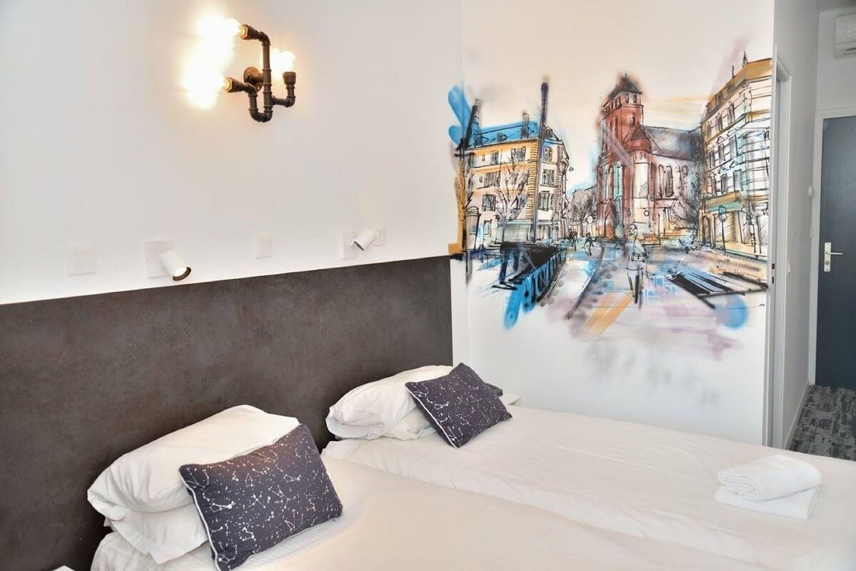A hotel room with a bed, two pillows, a wall-mounted light fixture, and a large mural of a city street.