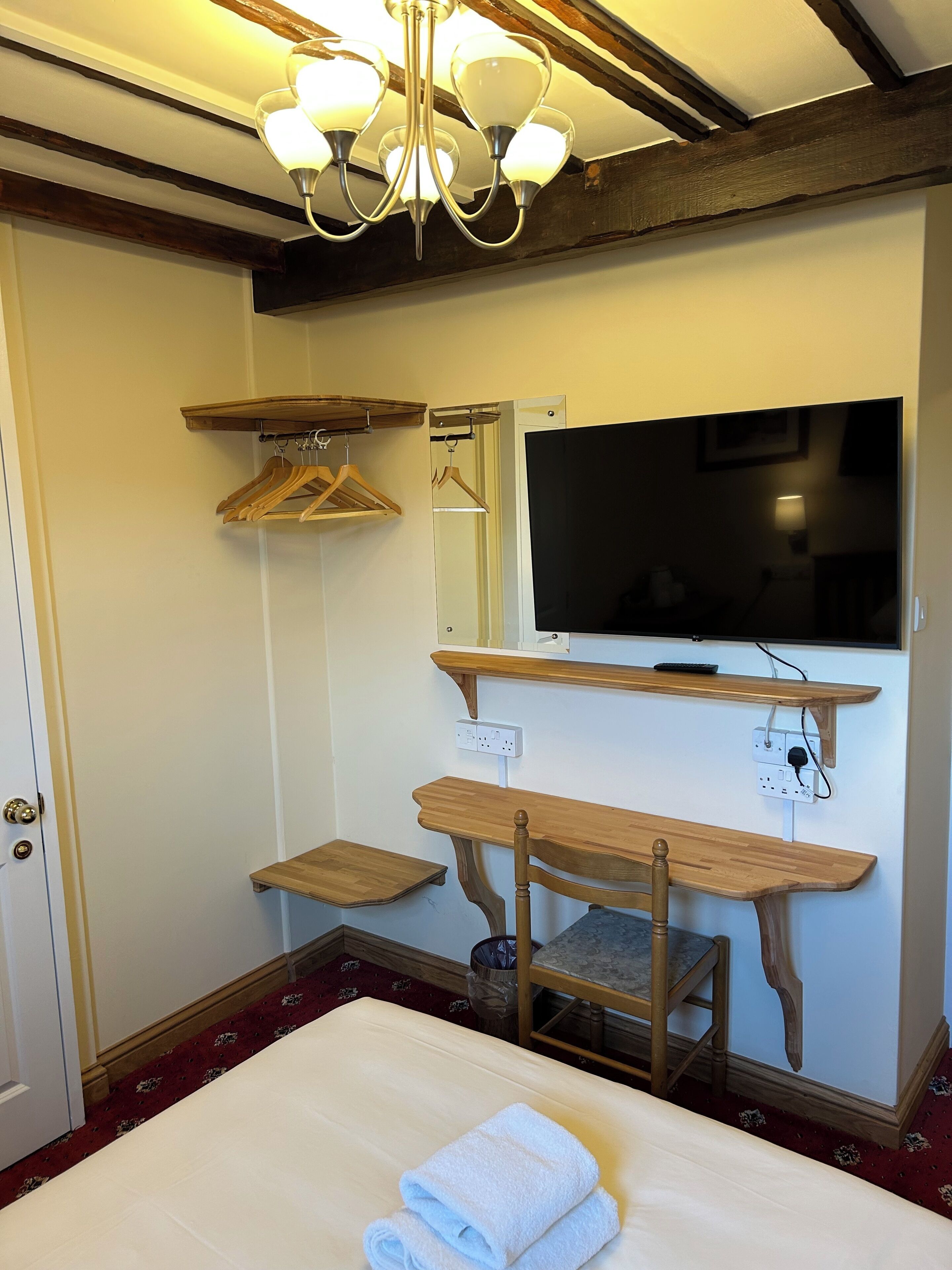 Quarto de hotel com cama, mesa de cabeceira de madeira, televisão fixada na parede, espelho e lustre.