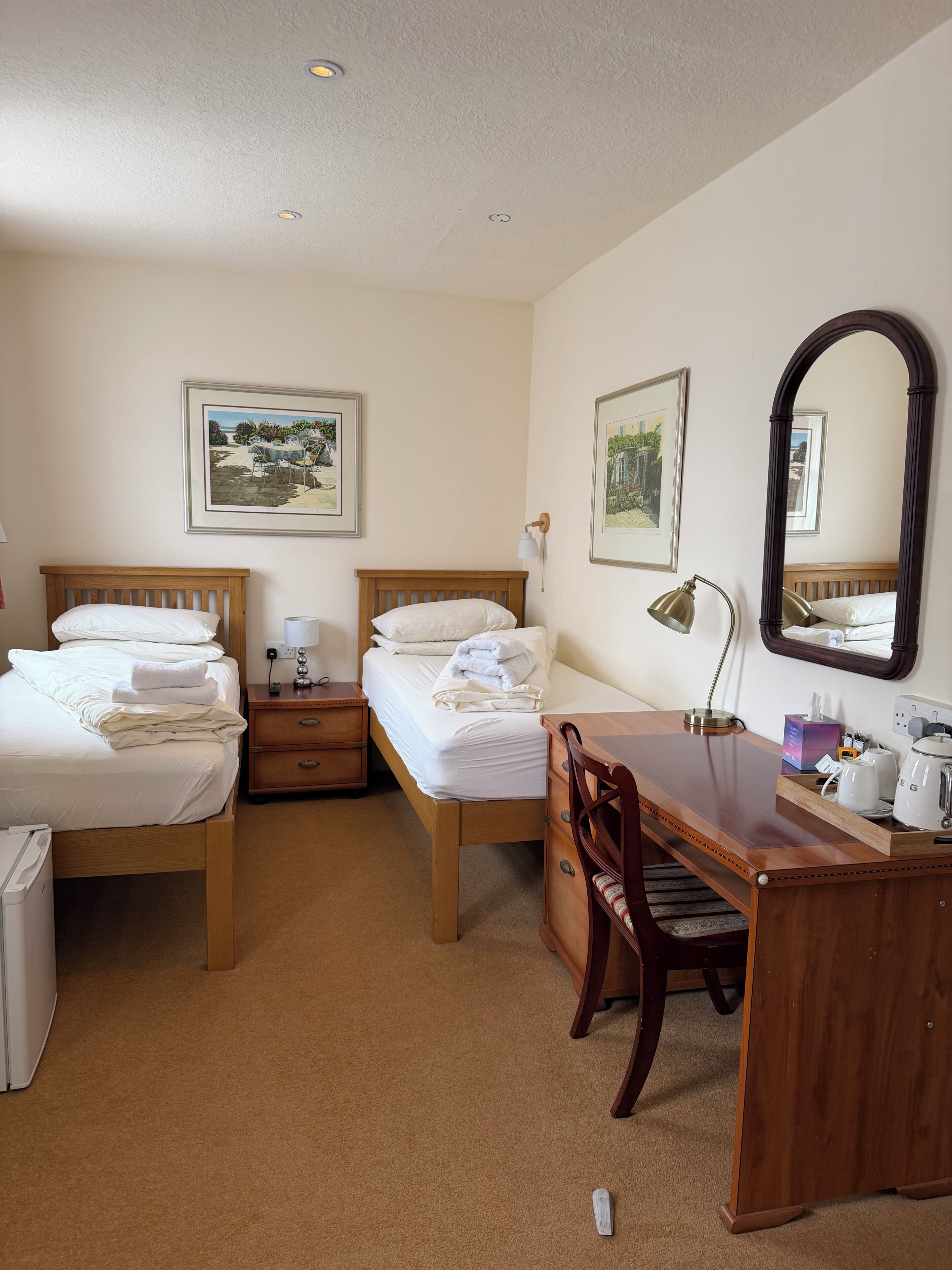 A hotel room with two beds, a desk, a chair, a mirror, and two wall-mounted lamps.