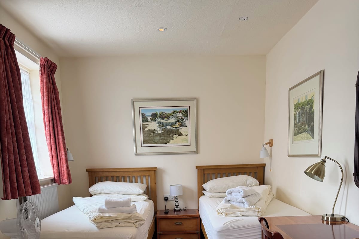 A hotel room with two single beds, a desk, a chair, a small refrigerator, a bedside table with a lamp, and two framed pictures on the wall.