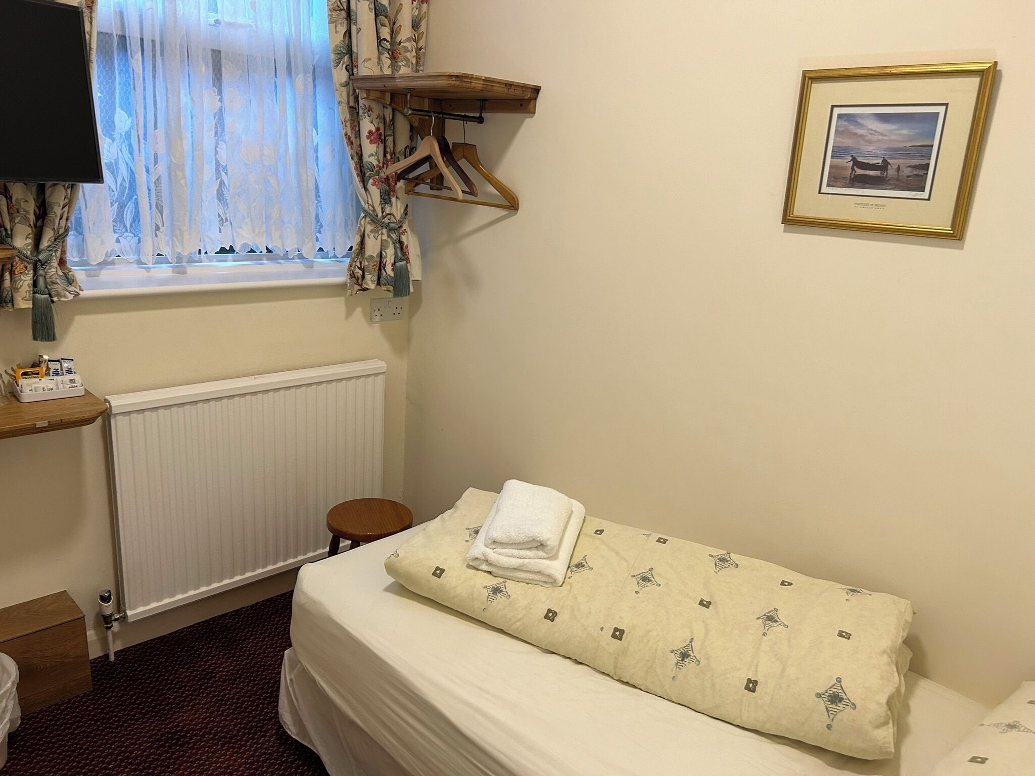 A hotel room with a bed, a bedside table with a lamp, a window with curtains, a radiator, and a framed picture on the wall.