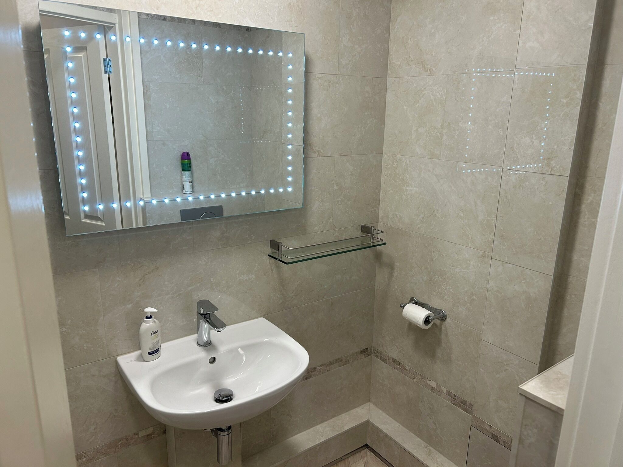A bathroom with a vanity sink, a mirror with LED lights, a glass shelf, and a toilet paper holder.