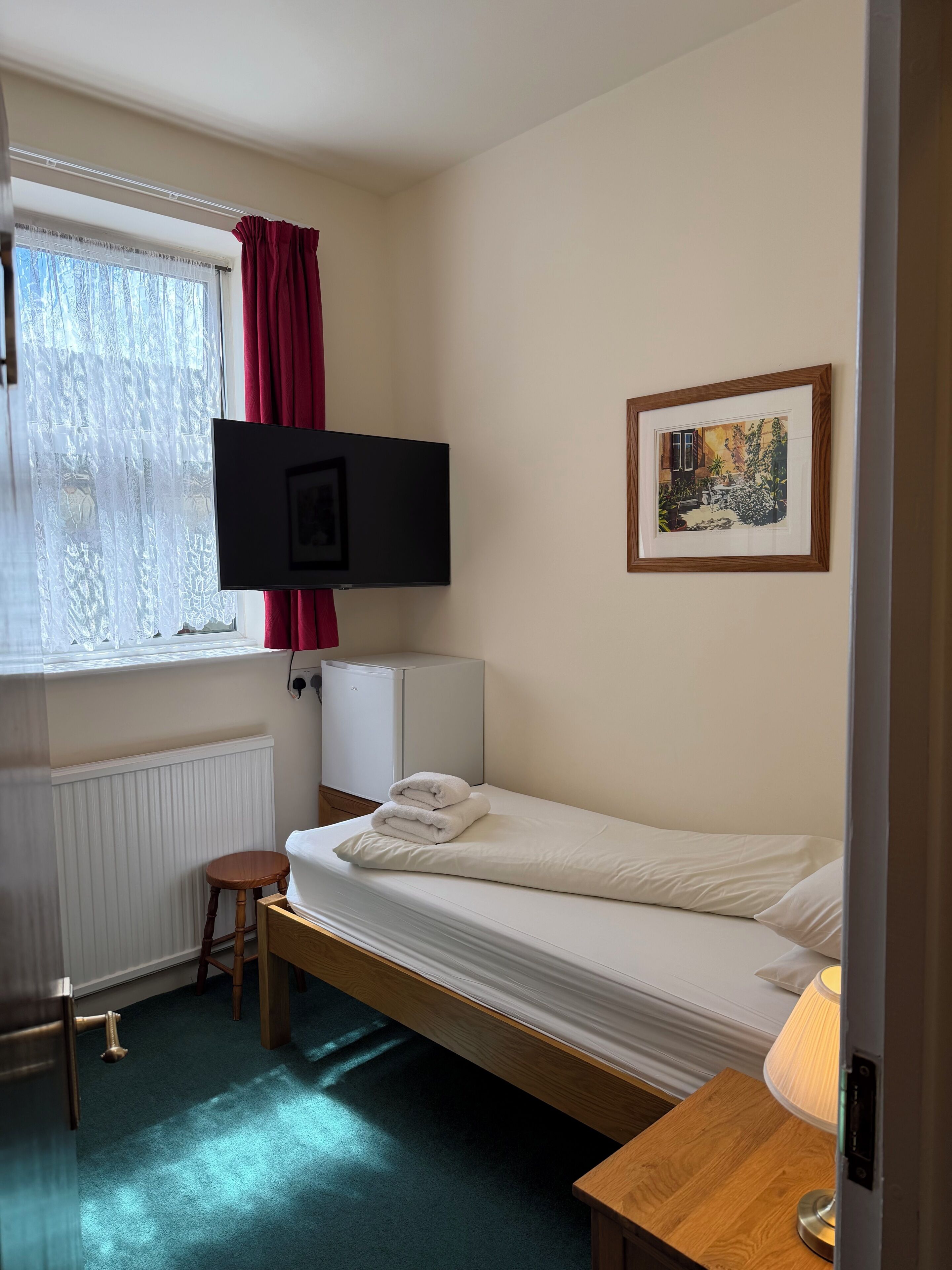 A single bed with a white mattress and pillows, a small wooden stool, a bedside table with a lamp, a TV mounted on the wall, and a radiator.