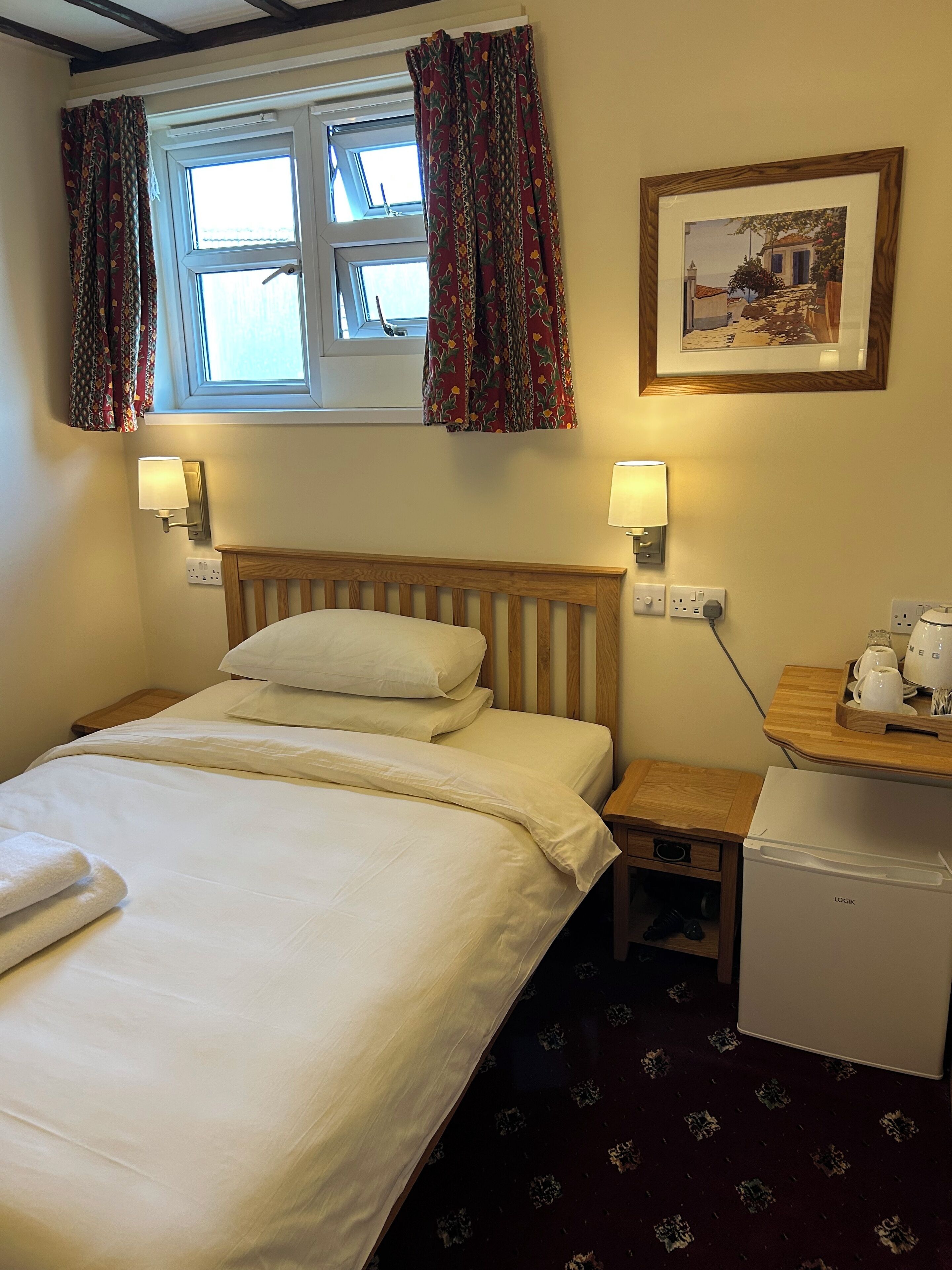 A hotel room with a bed, a small table with a kettle, a refrigerator, and a wall-mounted picture.