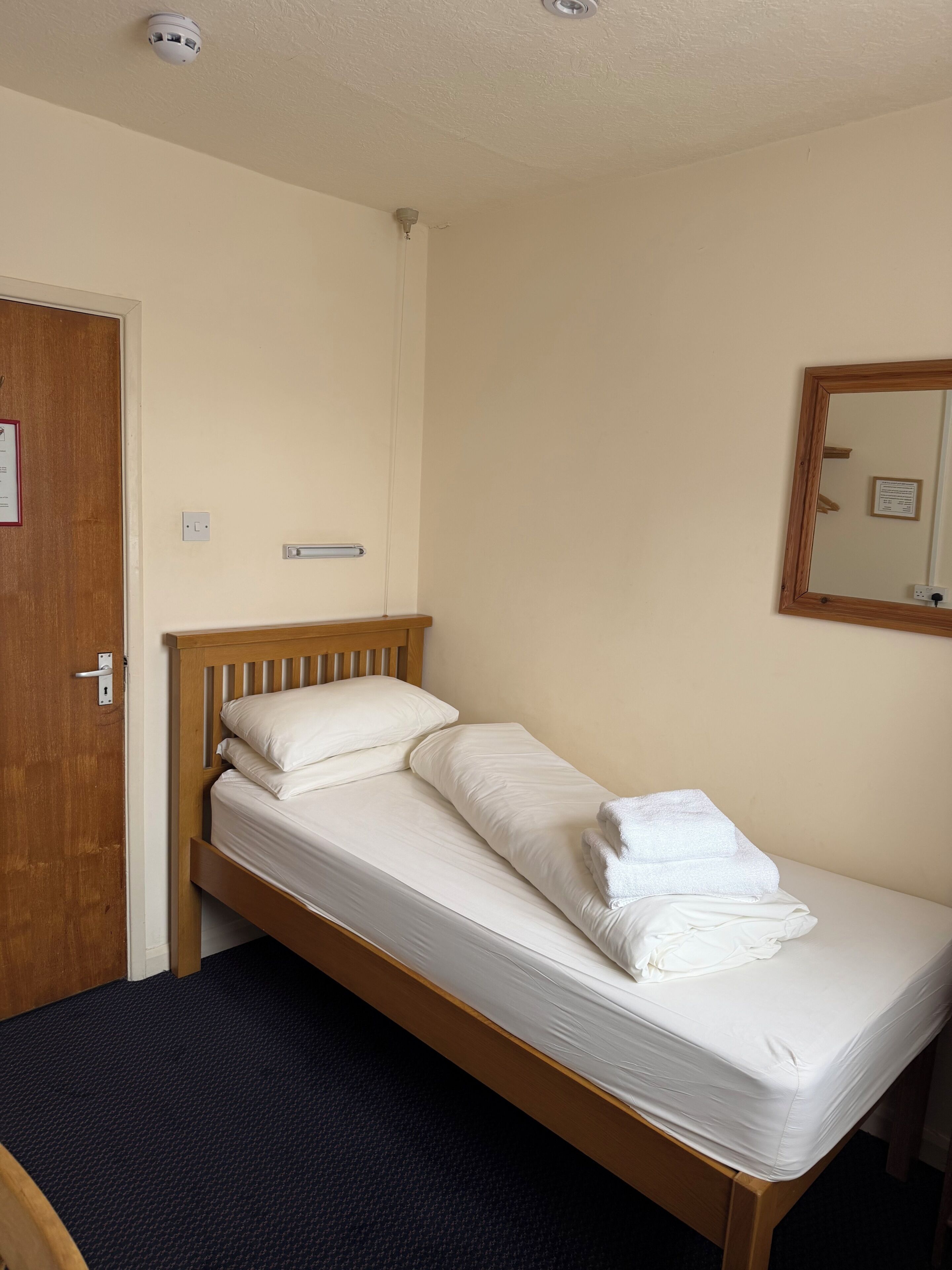 Una cama individual con ropa blanca, cabecera de madera, suelo cubierto de moqueta oscura, una puerta de madera y un espejo fijado a la pared.