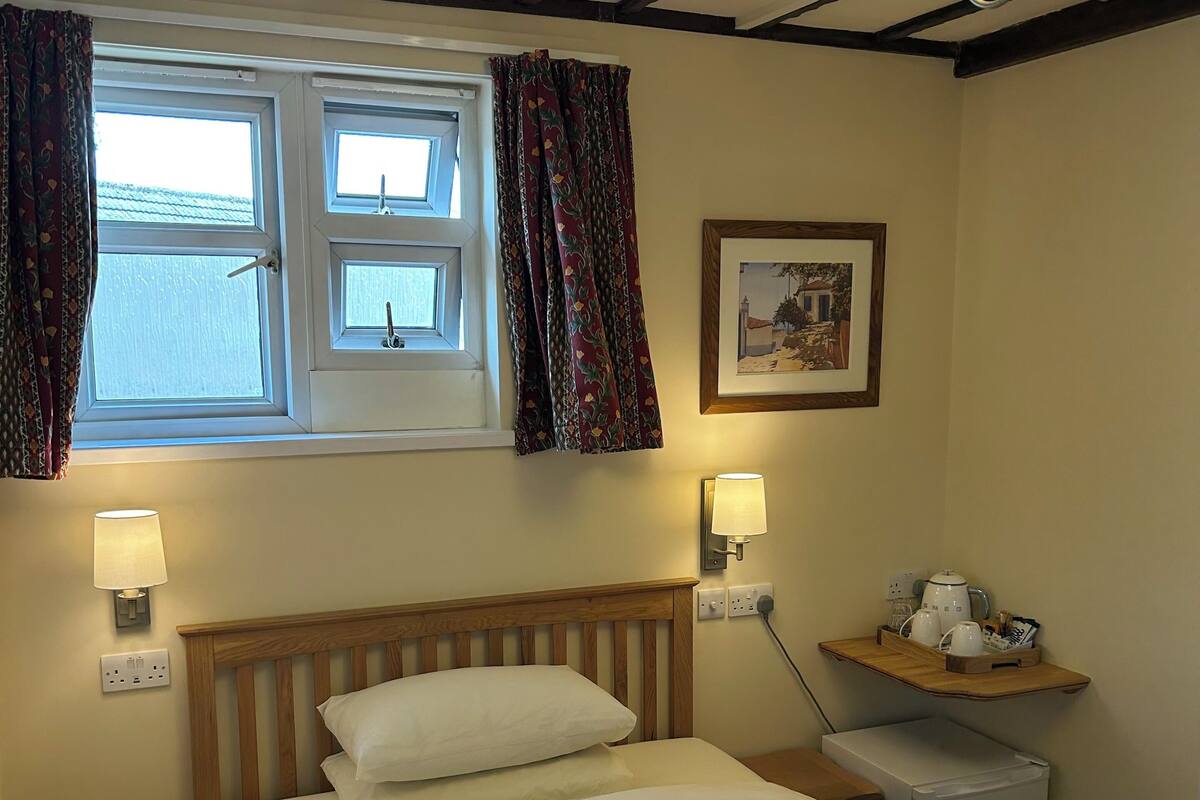 A neatly made bed with white linens, a bedside table with a lamp, a small refrigerator, and a shelf with a kettle and cups.