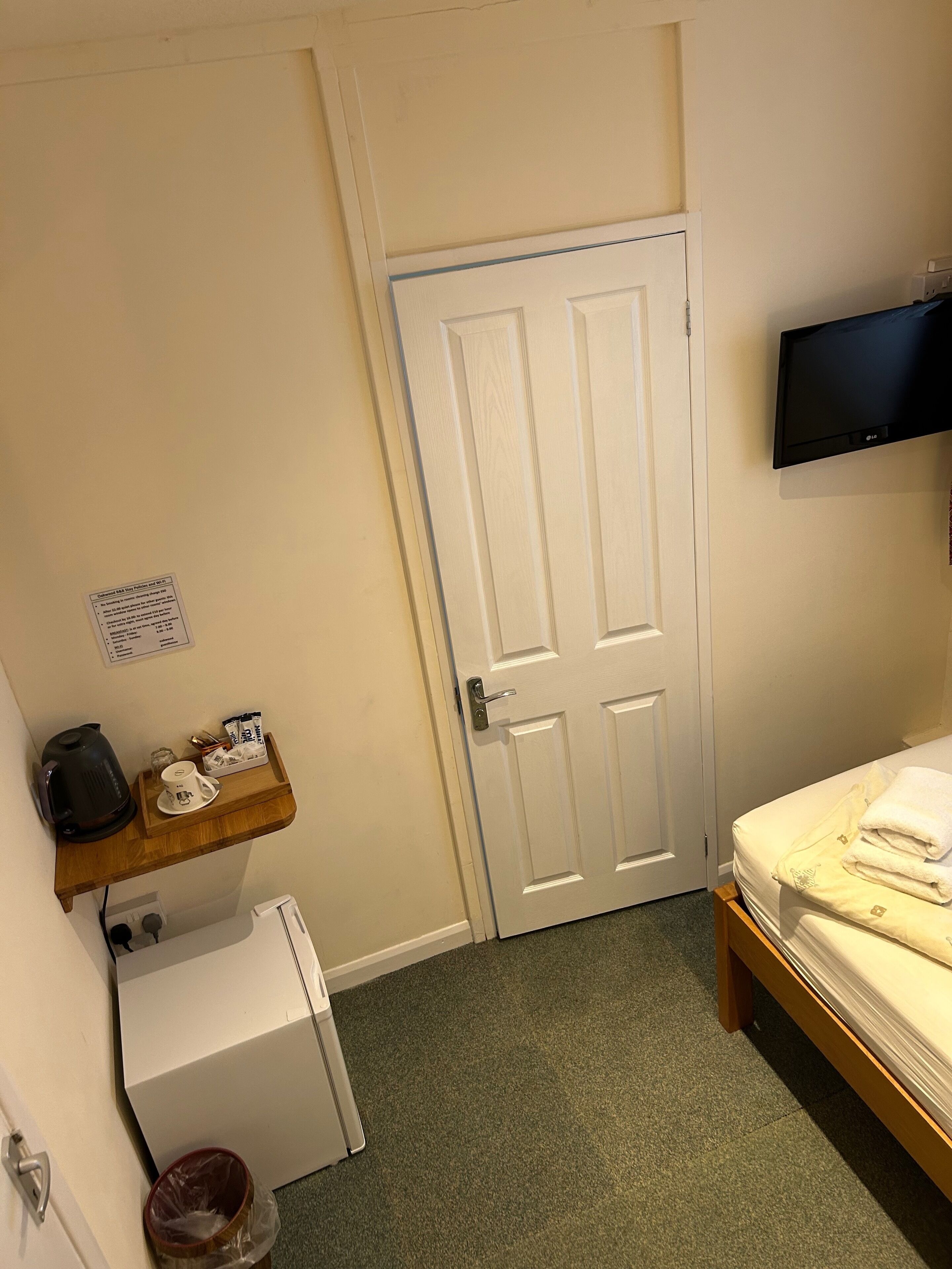 A small hotel room with a white door, a television mounted on the wall, a bed with white linens, a small wooden shelf with a kettle and a cup, and a white refrigerator.