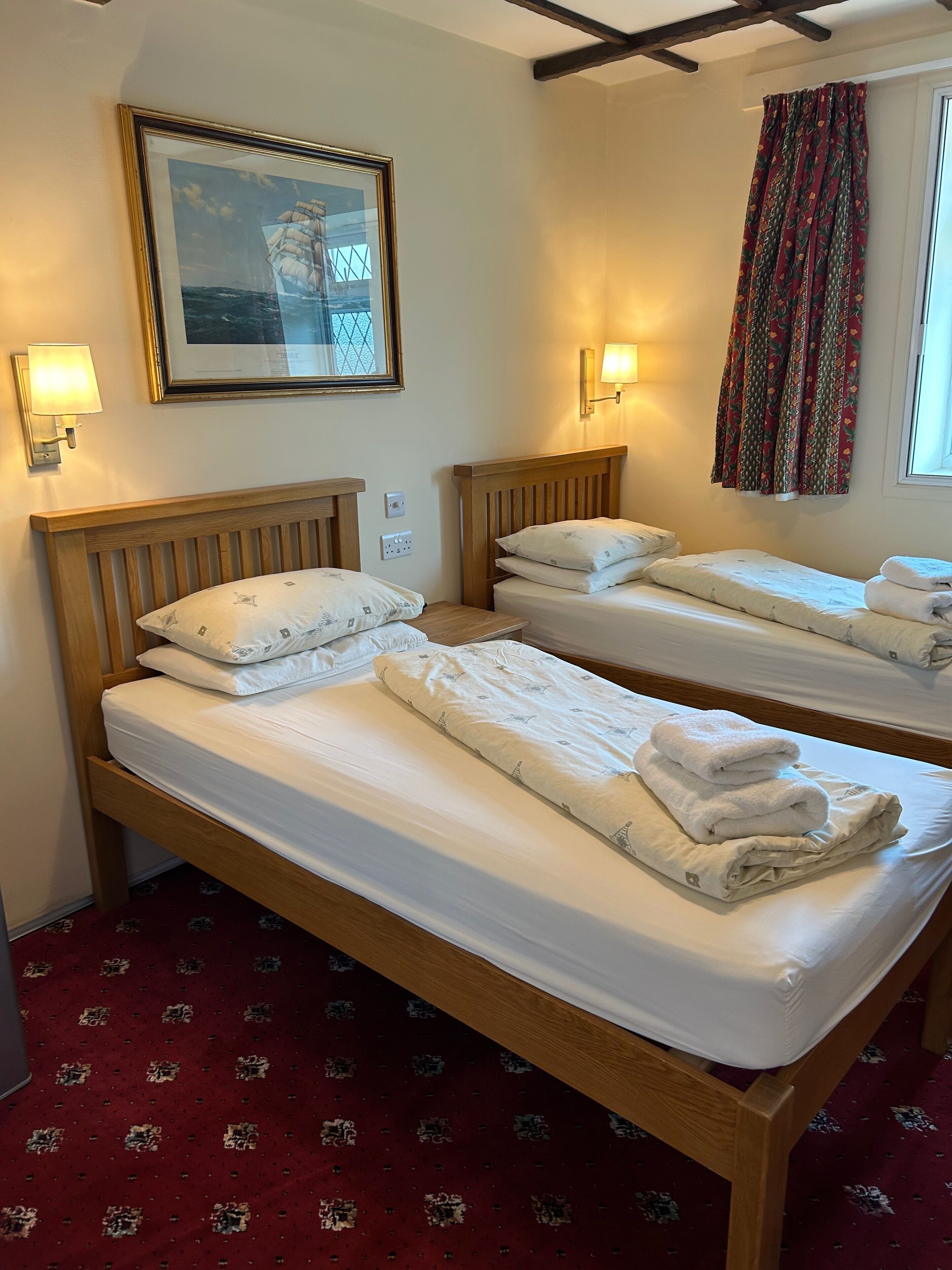 A hotel room with two wooden beds, each with white linens and pillows, a framed picture on the wall, and a window with patterned curtains.