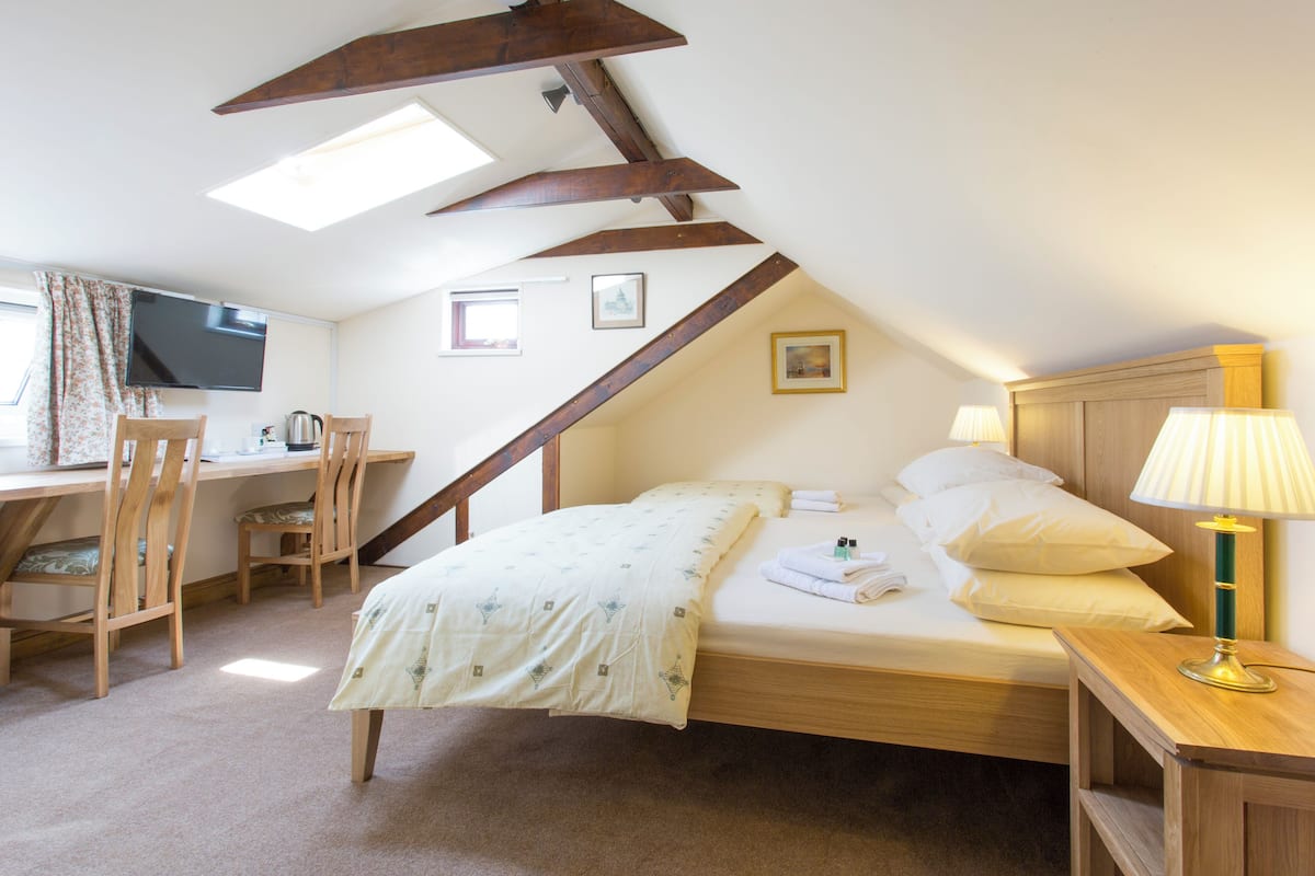 A bedroom with a bed, bedside table, lamp, desk with chair, and a television.