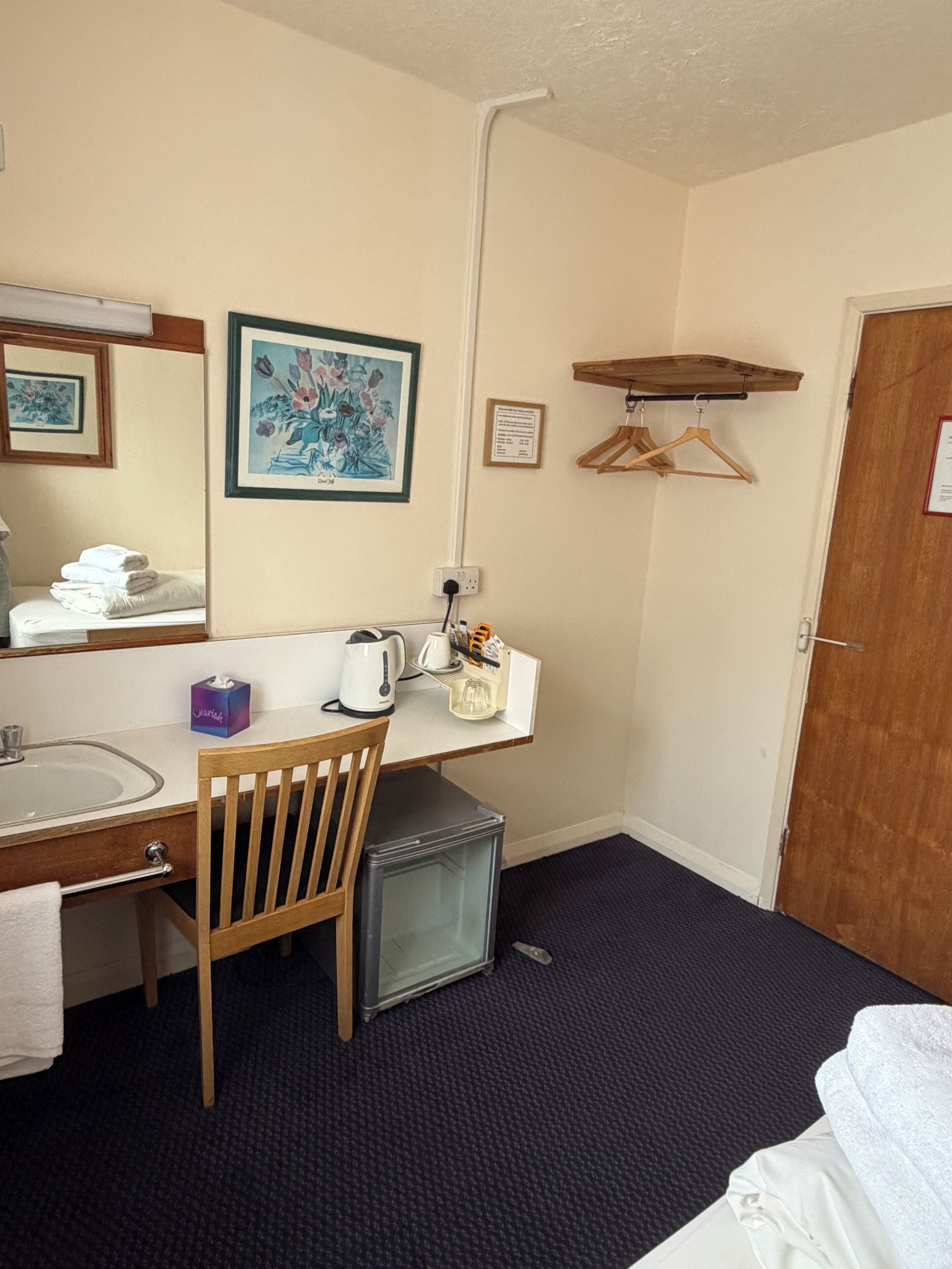 Une chambre d’hôtel avec une chaise en bois, un petit réfrigérateur, un lavabo, un miroir, une œuvre d’art florale encadrée, une étagère en bois avec des cintres et une porte.