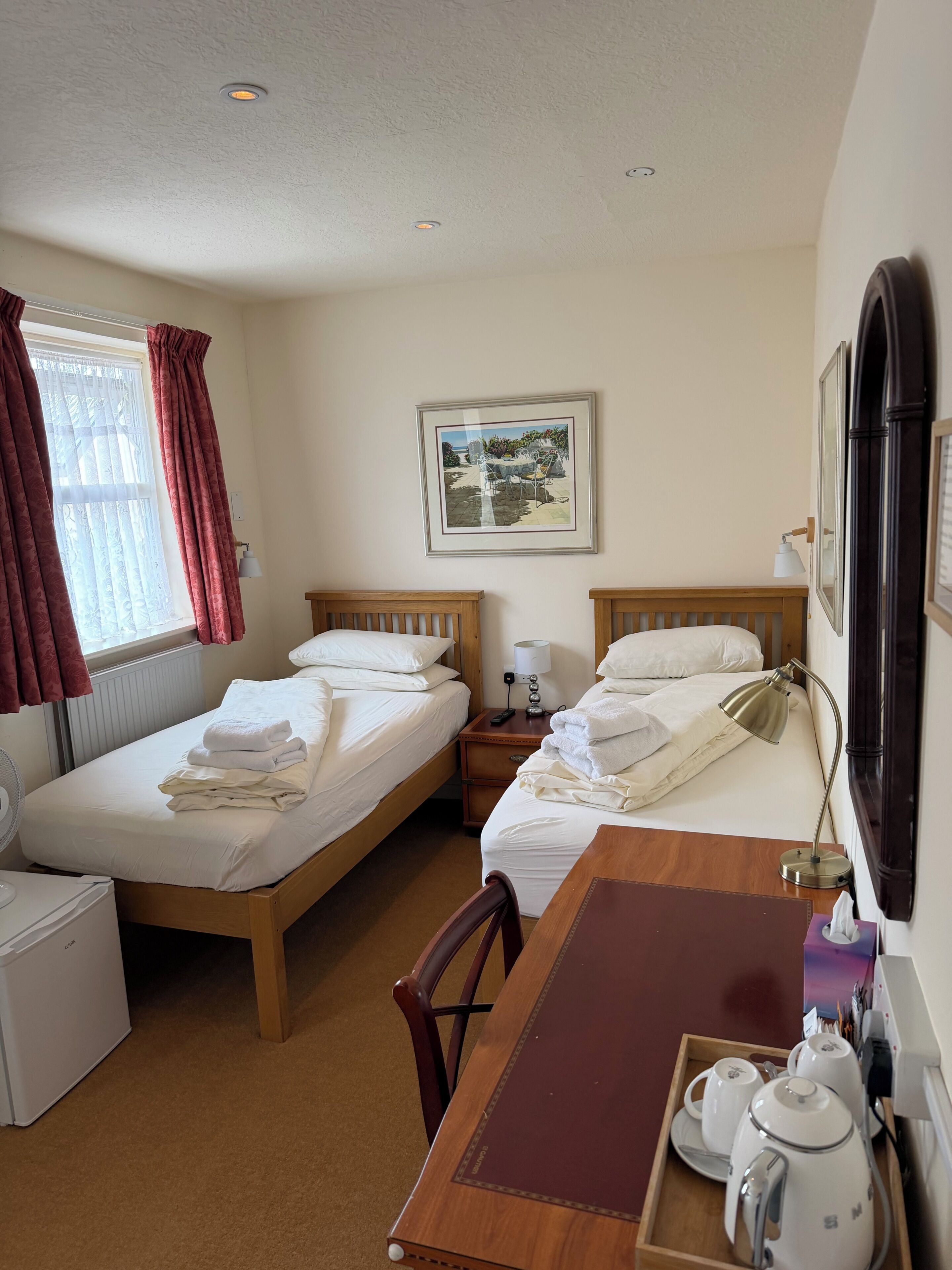 A hotel room with two beds, a wooden table with a tray of tea and cups, a desk lamp, and a framed picture on the wall.