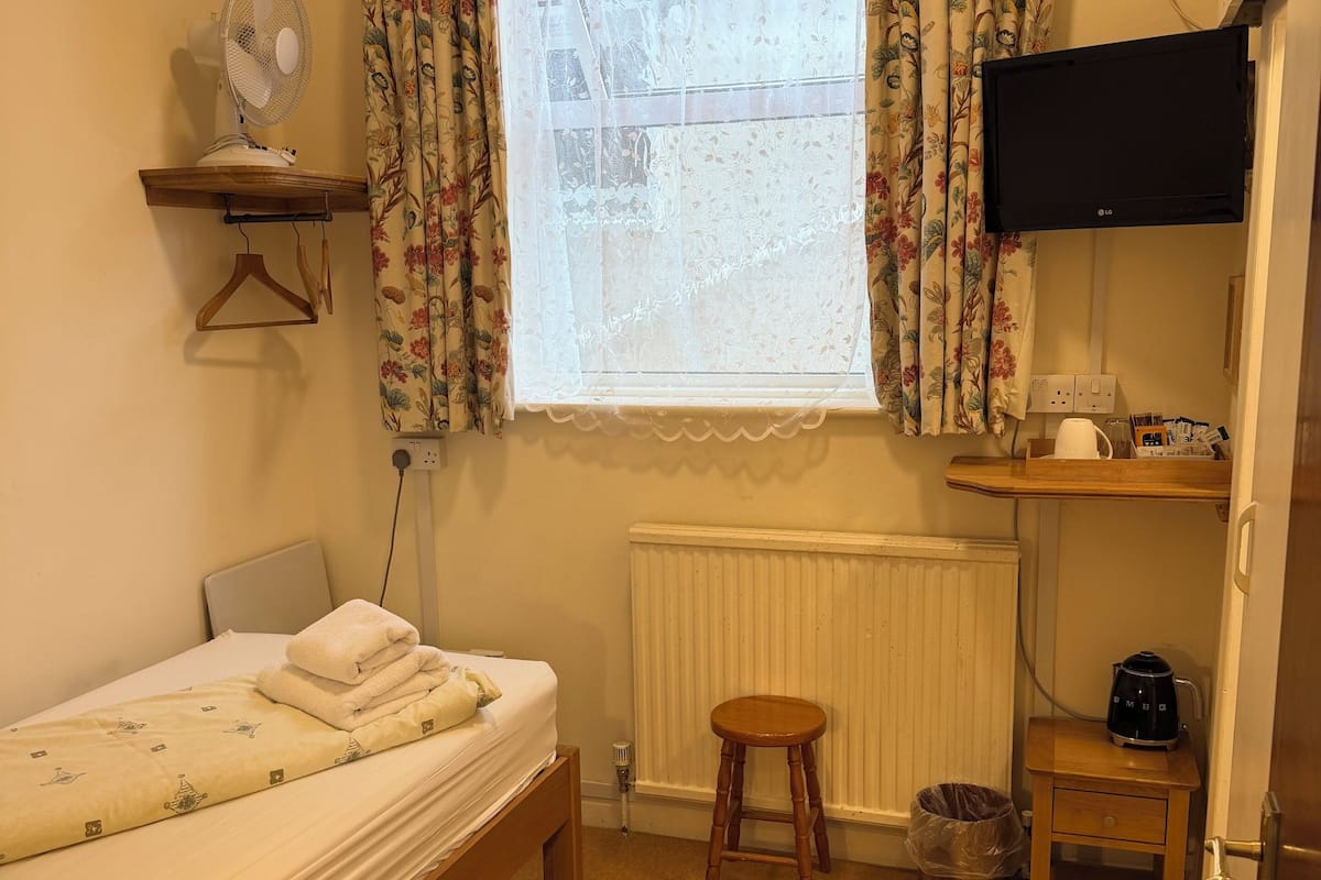 Une chambre à coucher avec un lit, un tabouret en bois, un radiateur, une télévision et une petite table sur laquelle se trouve une bouilloire.