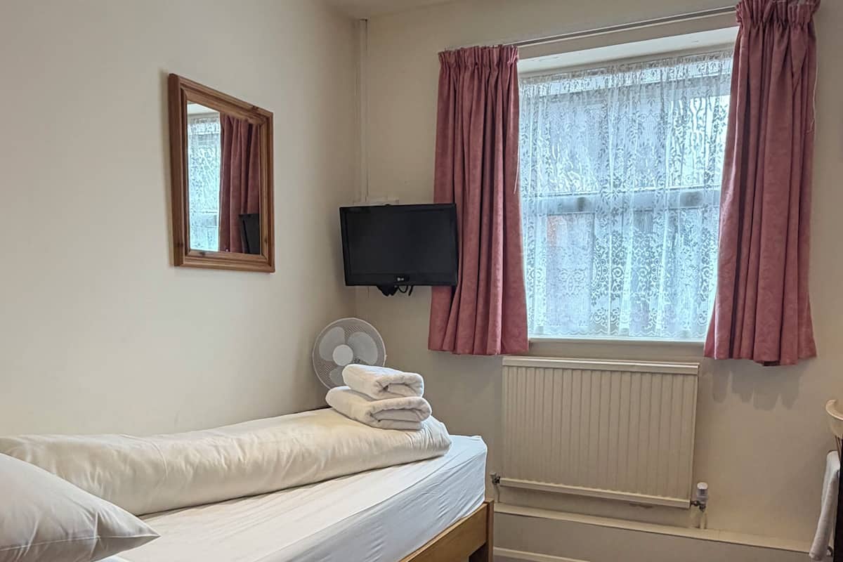 A single bed with white linens, a wall-mounted TV, a window with pink curtains, and a radiator.