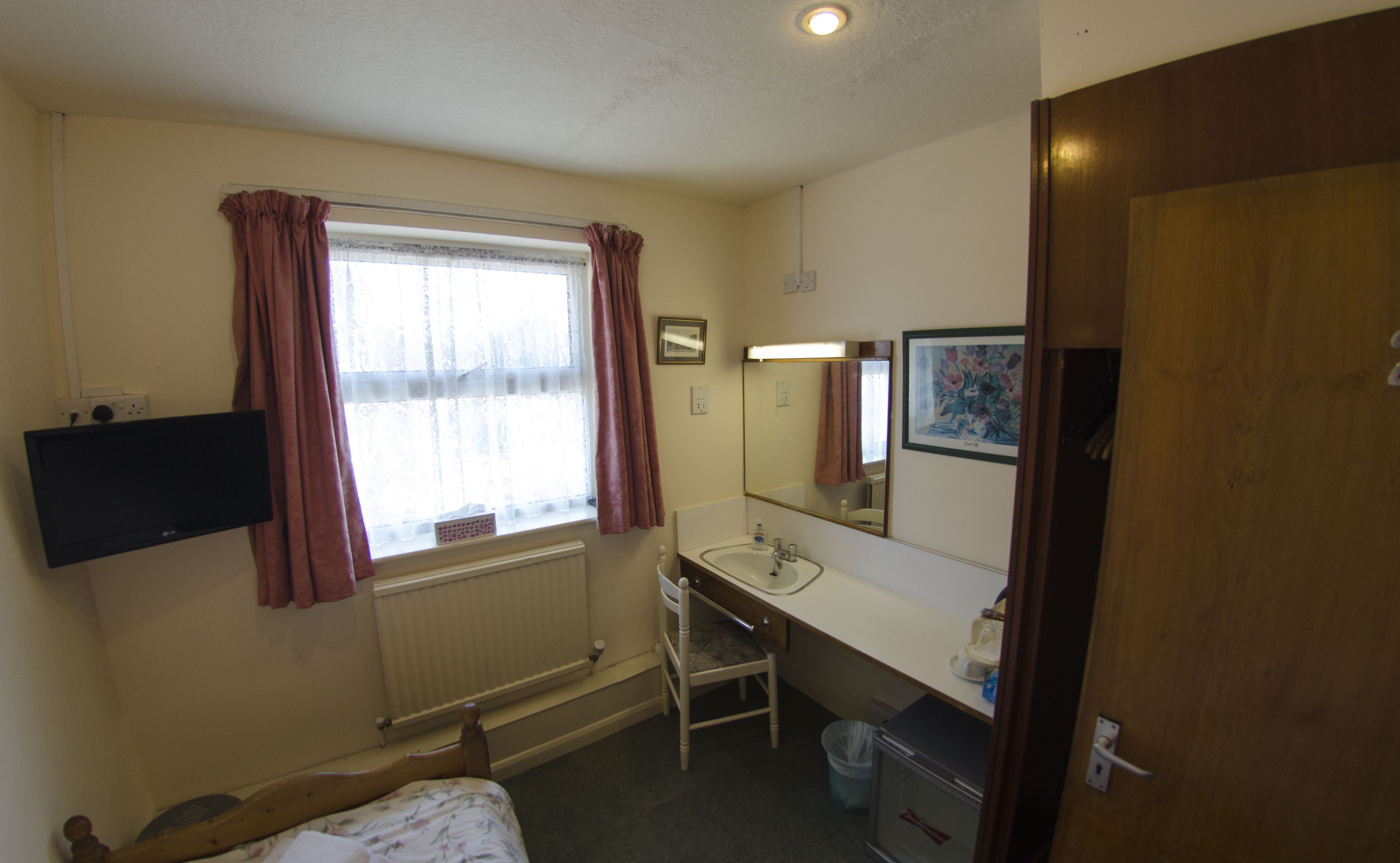 A small, well-lit room with a bed, a desk, a chair, a window with curtains, a mirror, and a radiator.