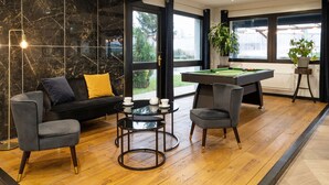 Una habitación moderna con mesa de billar, sofá y dos sillones. Se aprecia un ventanal con vistas a la vegetación.