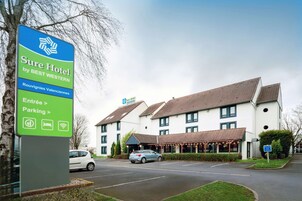 Señal de hotel que indica "Sure Hotel by BEST WESTERN" con indicaciones a "Entrada > Estacionamiento >" y símbolos para Wi-Fi y estacionamiento.