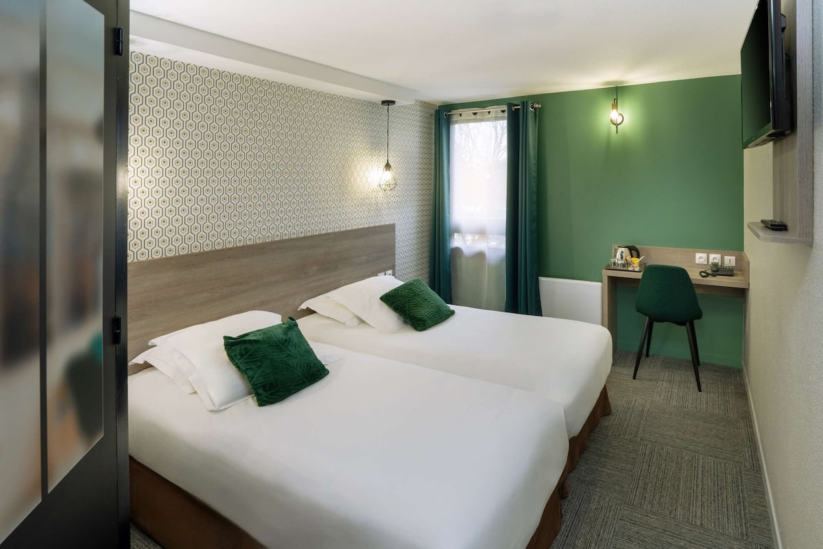 A hotel room with two beds, a desk, a chair, a TV, and a patterned wall.