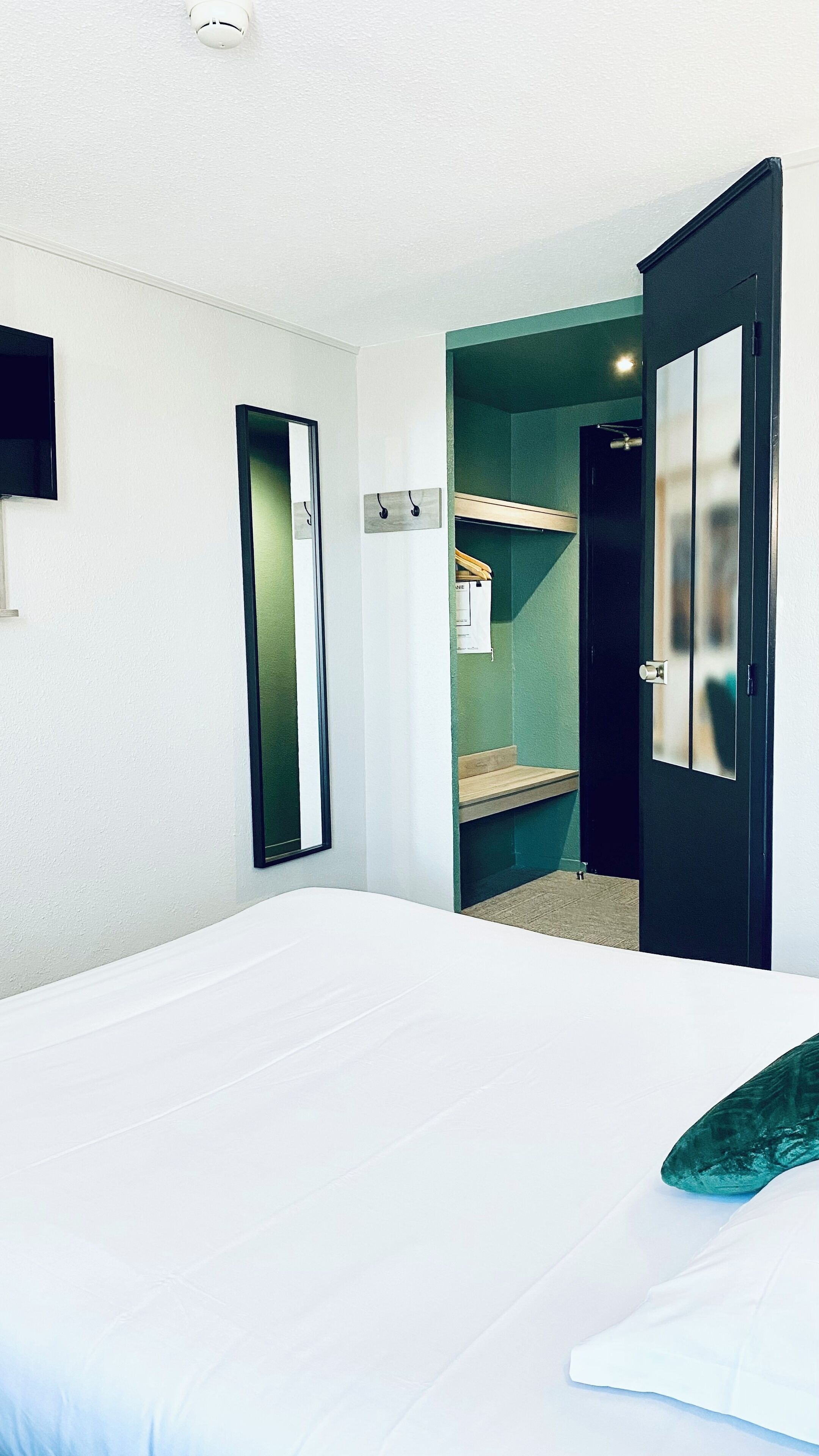 A hotel room with a large bed, a mirror, a door leading to a bathroom, and a small shelf with toiletries.