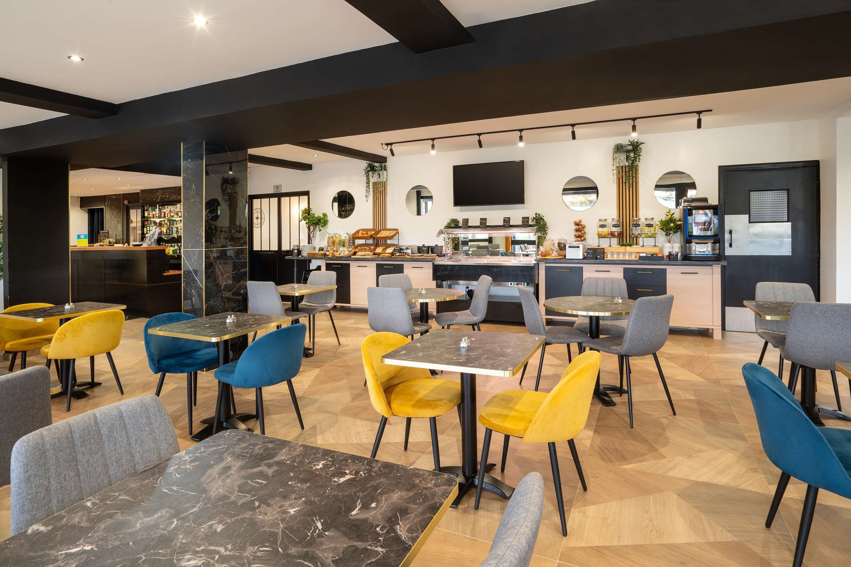 A modern restaurant with marble tables and colorful chairs, a kitchen area, and a flat-screen TV on the wall.