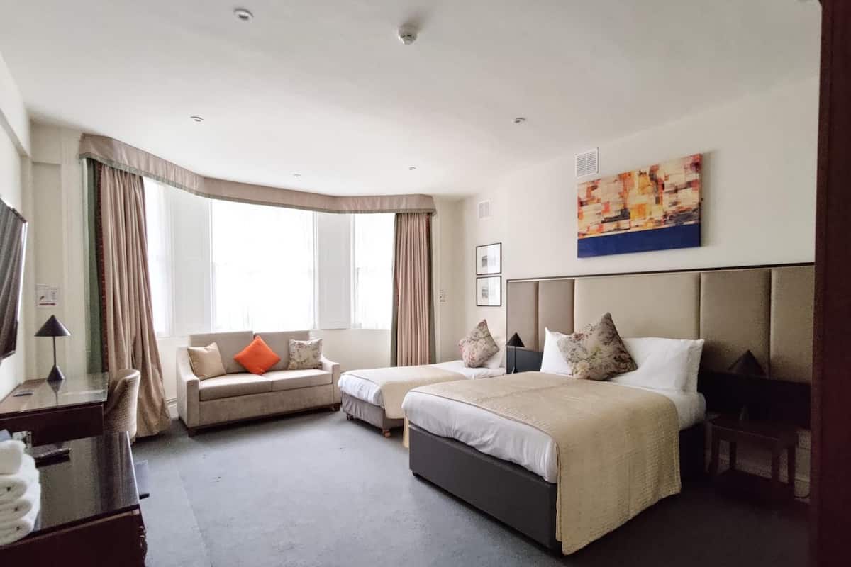 A hotel room with a sofa, two beds, a desk, and a large window.