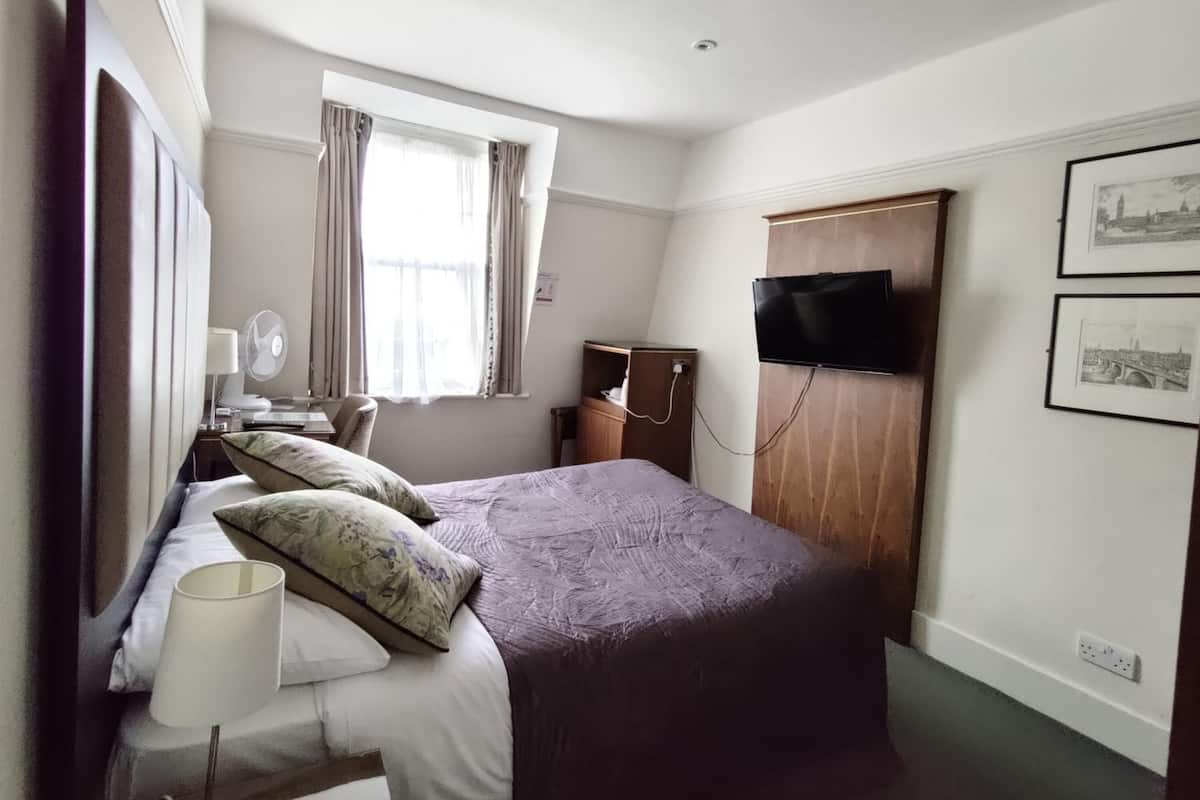 A hotel room with a large bed, a desk with a lamp, a TV mounted on the wall, and a window with curtains.