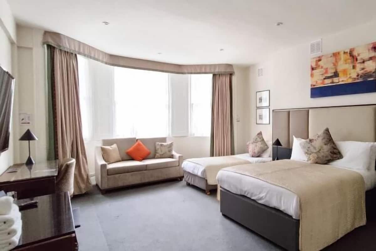 A hotel room with a curved window, a sofa, a bed with a headboard, and a painting on the wall.