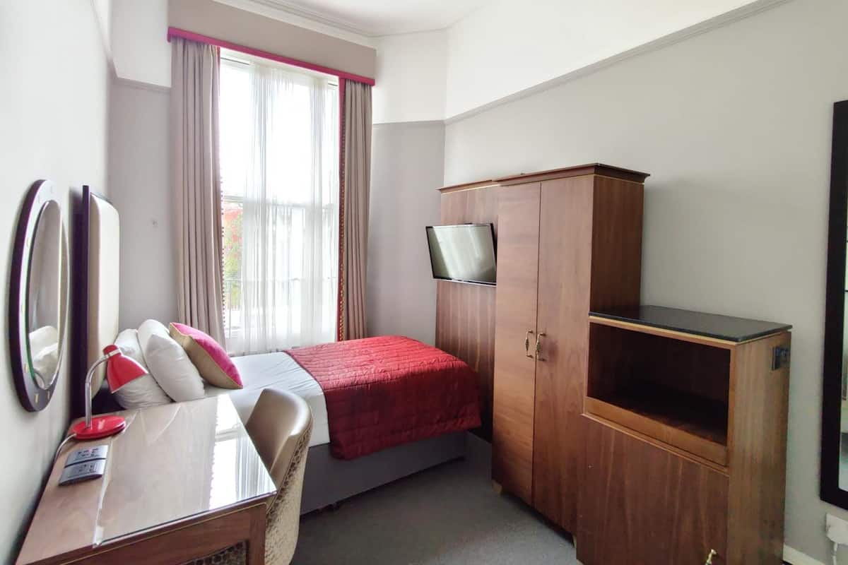 A small, neatly arranged hotel room with a bed, a desk with a chair, a television mounted on a wooden cabinet, and a window with curtains.