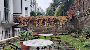 A courtyard with a round table and benches, surrounded by brick walls and a variety of plants.