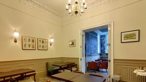 A dining area with wooden tables and chairs, a green cushioned bench, and a chandelier. There are framed botanical prints on the walls and a radiator on the right.