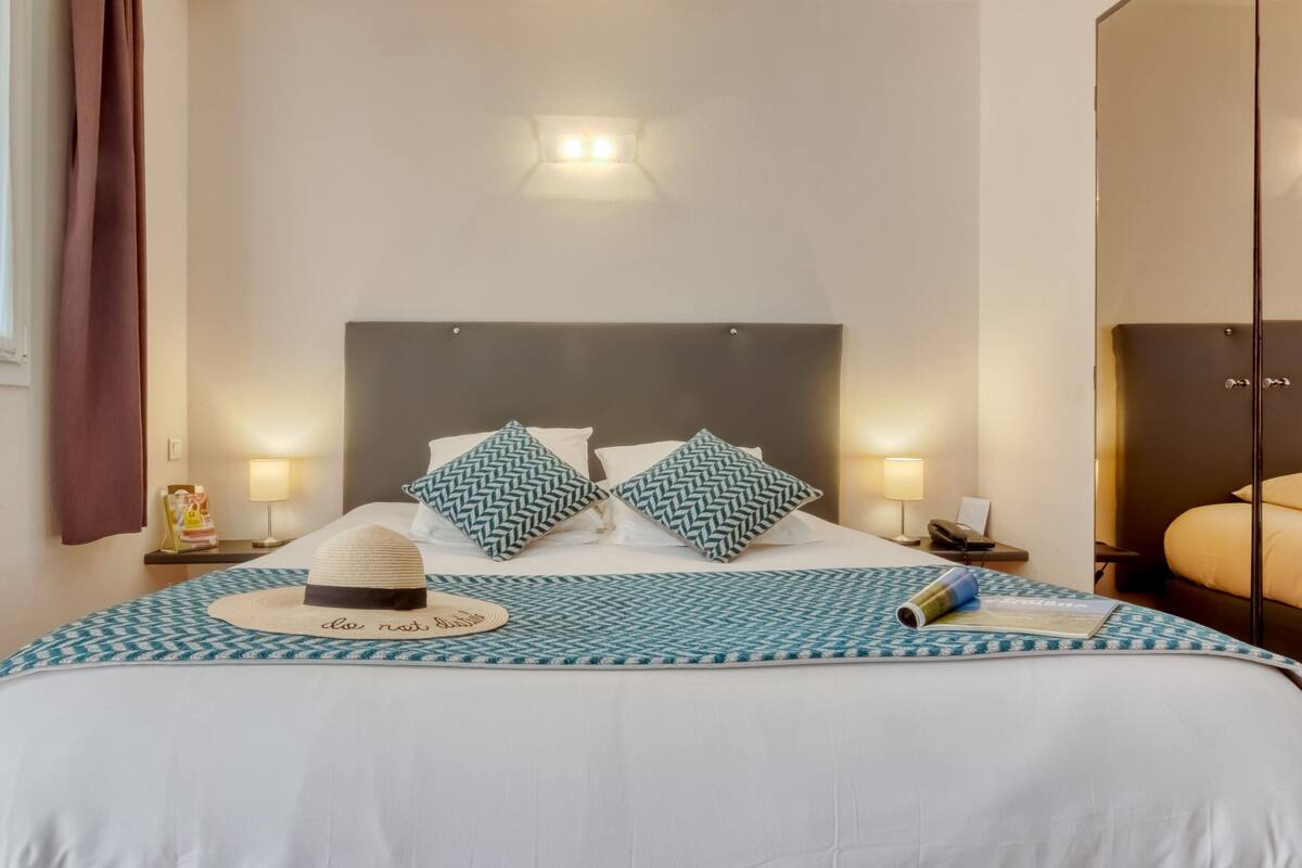 A neatly made bed with a patterned bedspread, two decorative pillows, a hat, and a book on the nightstand.