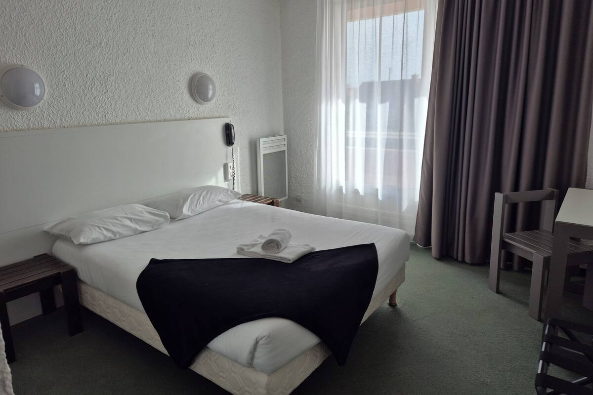 Quarto de hotel com cama, escrivaninha, cadeira, telefone e janela com cortinas.
