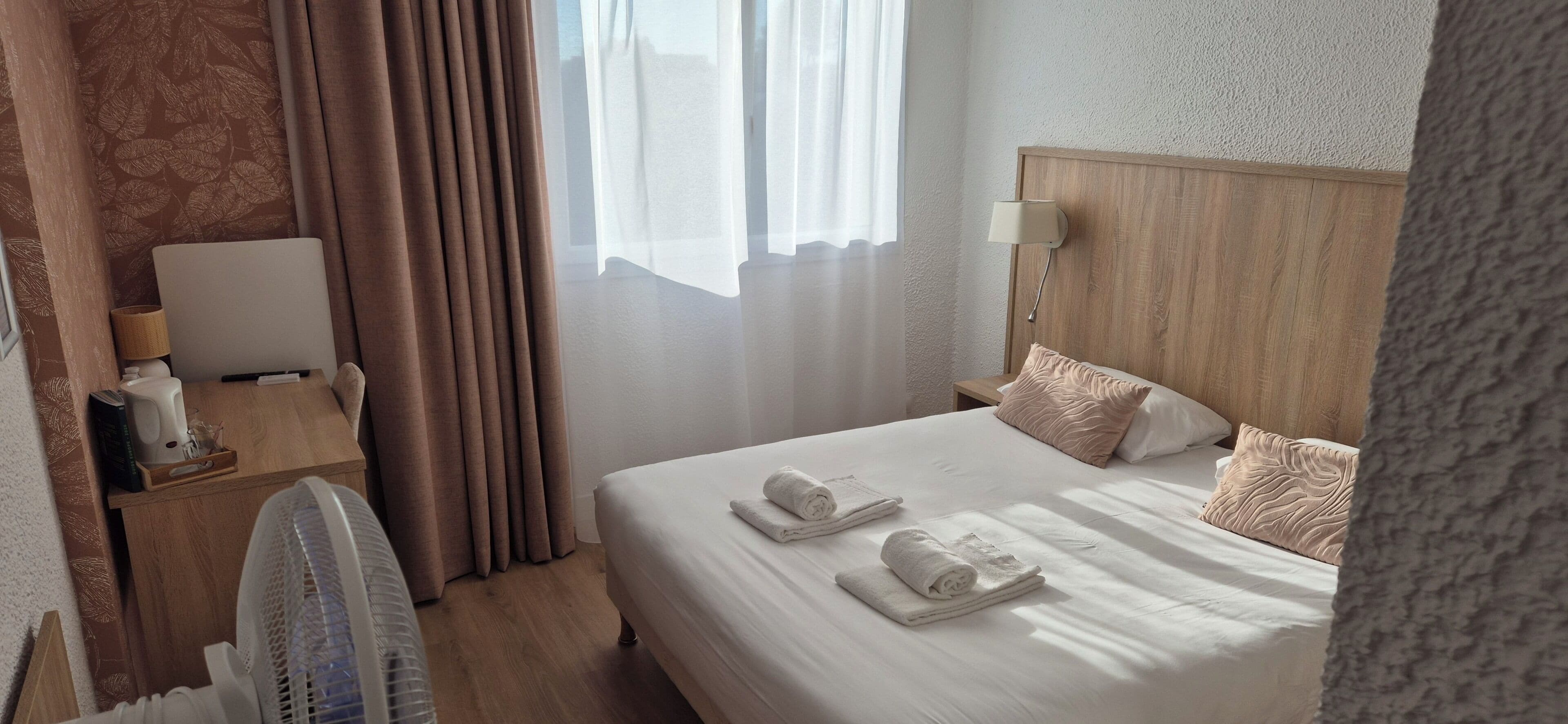A hotel room with a bed, a bedside table with a lamp, a fan, and a window with curtains.