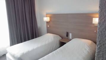 Quarto de hotel com duas camas de solteiro, cabeceira de madeira, abajures e uma janela com cortinas.