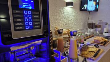 En kaffemaskine med digitalt display, et vægmonteret fjernsyn og et morgenmadsbord med brød, kager og tilbehør.