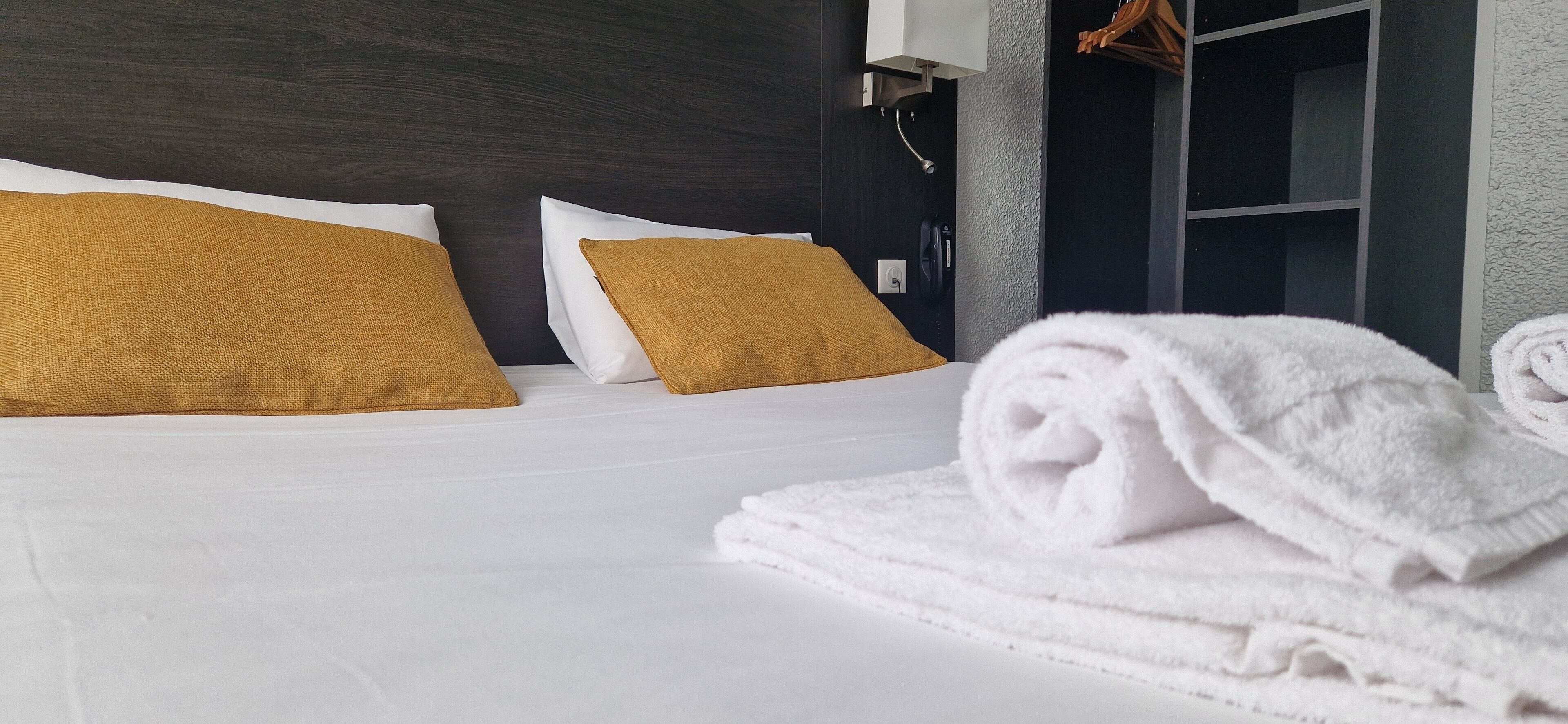 Een hotelkamer met een bed, witte en mosterdgele kussens, en een keurig opgevouwen witte handdoek.