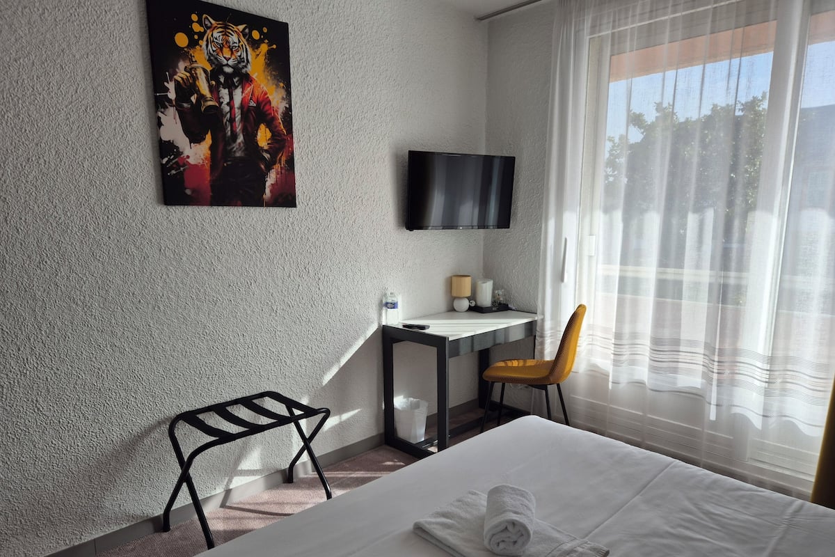 Quarto de hotel com cama, escrivaninha, cadeira, televisão e um quadro na parede.