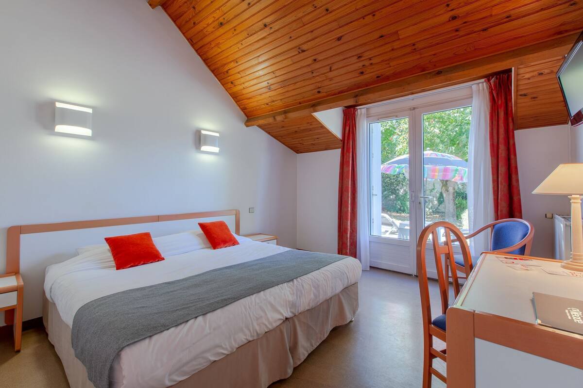 A bedroom with a wooden ceiling, a bed with red pillows, a desk with a chair, and a window with curtains.