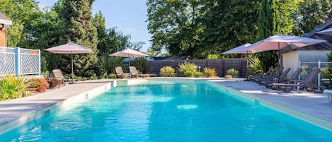 A rectangular swimming pool with a wooden deck, surrounded by lounge chairs and umbrellas, with a fence and trees in the background.