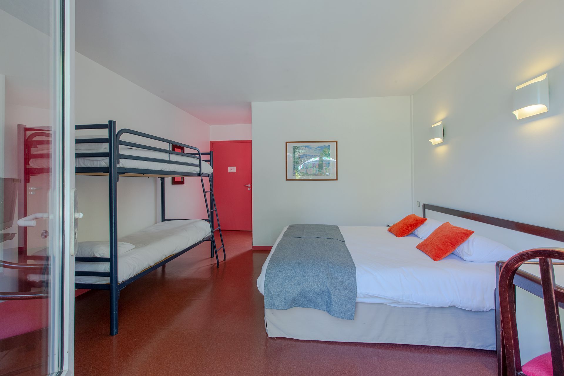 A dormitory room with bunk beds, a single bed with orange pillows, a framed picture on the wall, and a red door.