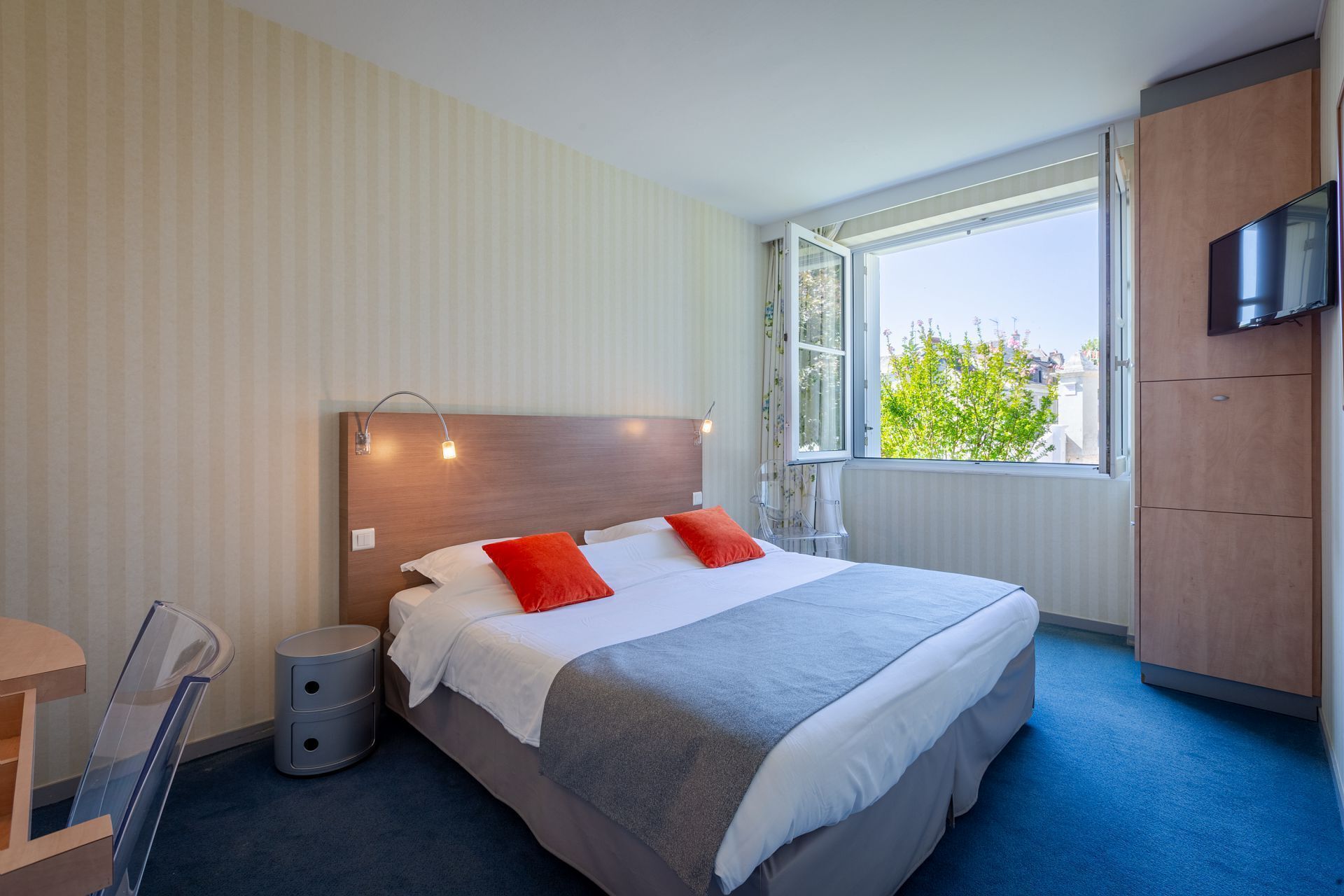 Une chambre d’hôtel avec un grand lit, une tête de lit en bois, une télévision, une fenêtre donnant sur un paysage verdoyant et un bureau avec une chaise.