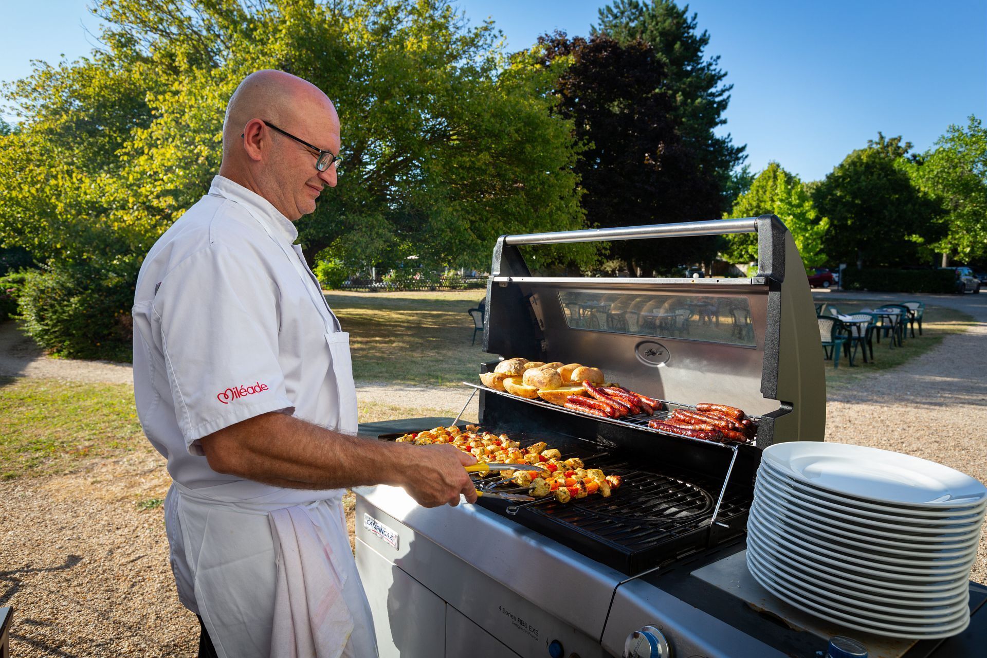 Un chef qui fait griller des saucisses et des brochettes sur un barbecue extérieur.