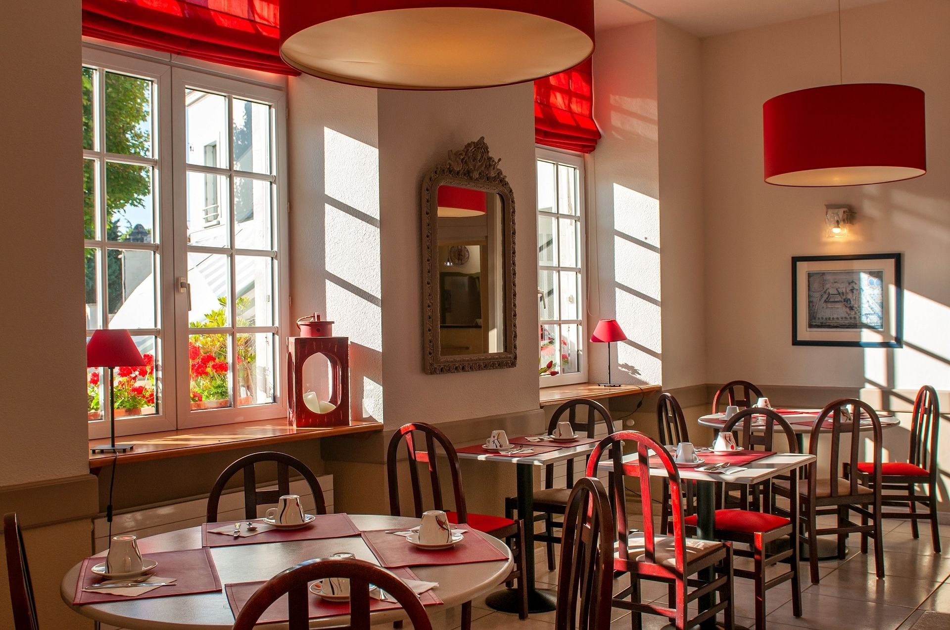 Un comedor con varias mesas y sillas, ventanas amplias con cortinas rojas y un espejo en la pared.