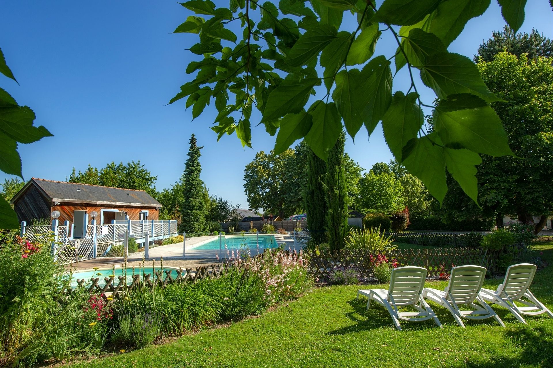 Un área de piscina con sillas de descanso, un edificio de madera con terraza cercada y follaje circundante.
